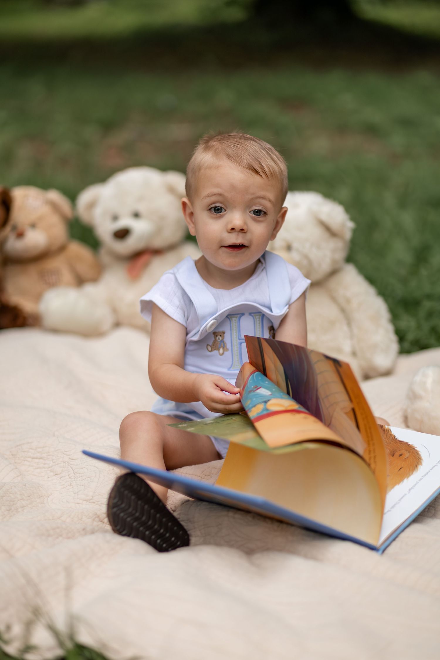 Toddler sitting on blanket outdoors reading colorful picture book surrounded by teddy bears in green garden setting.