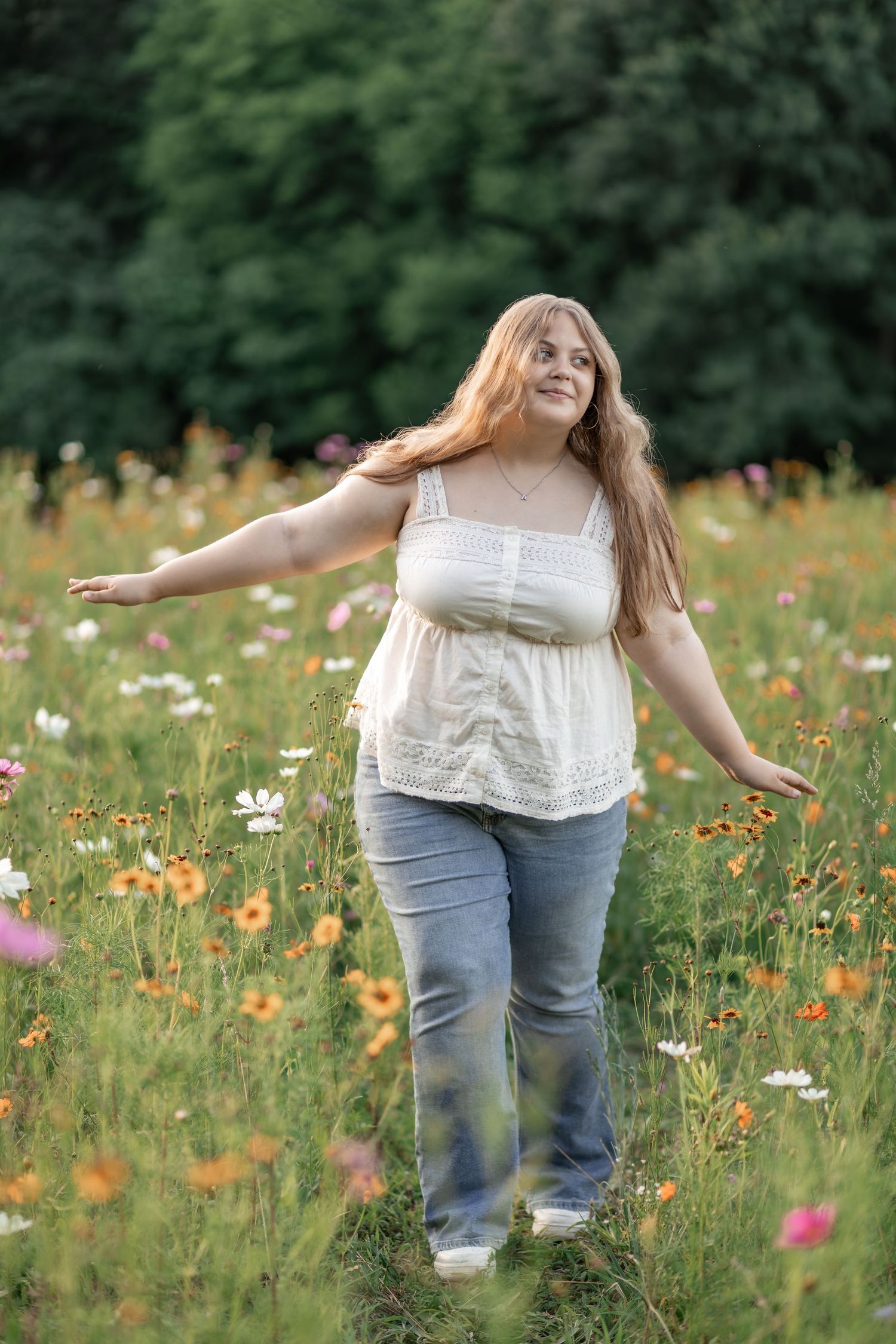 Young woman with long hair joyfully walking through a colorful wildflower meadow in jeans and white top.