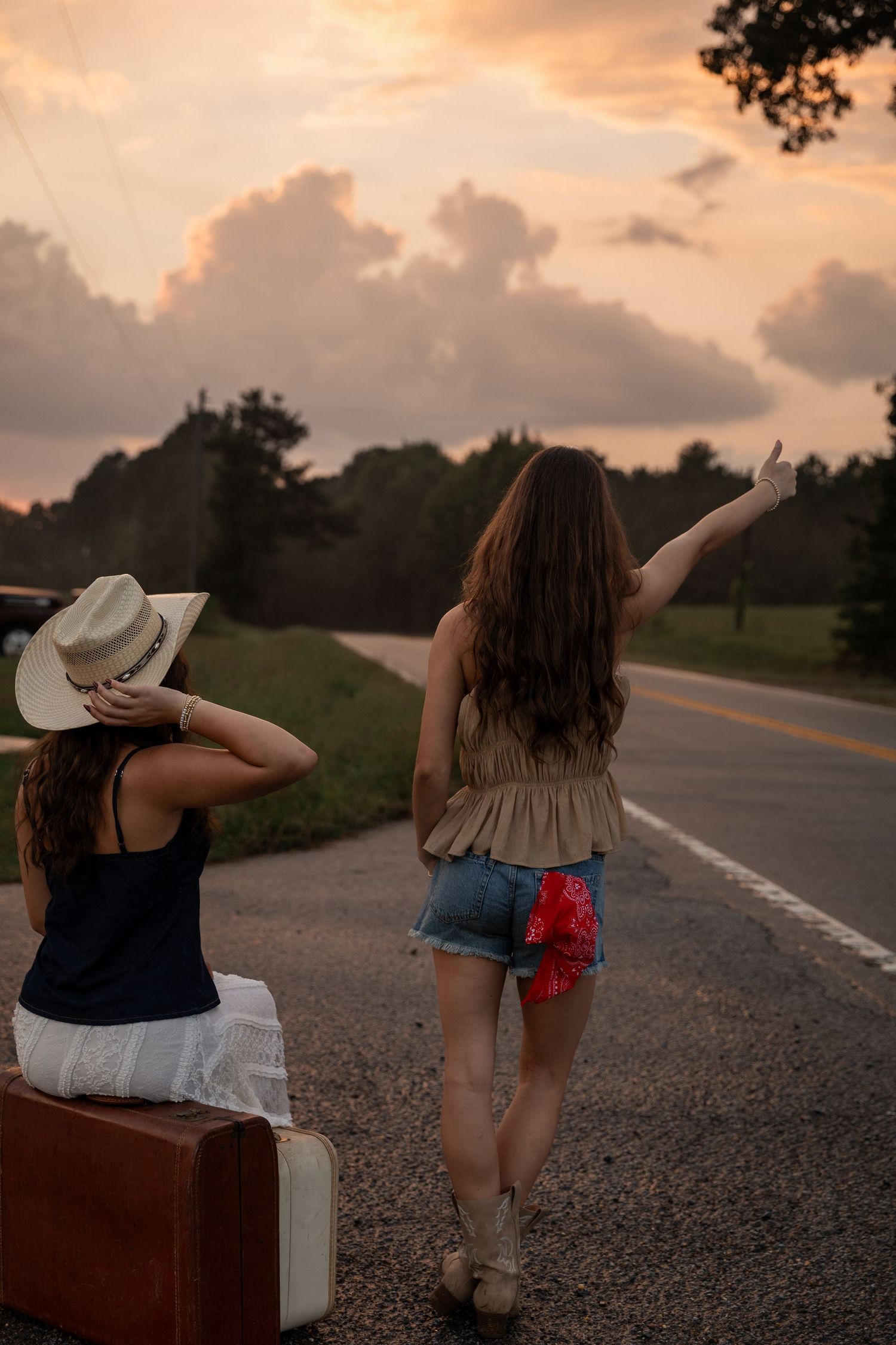 Two women hitchhiking at sunset on a rural road, one in cowboy hat sitting on vintage suitcase, other signaling passing cars.