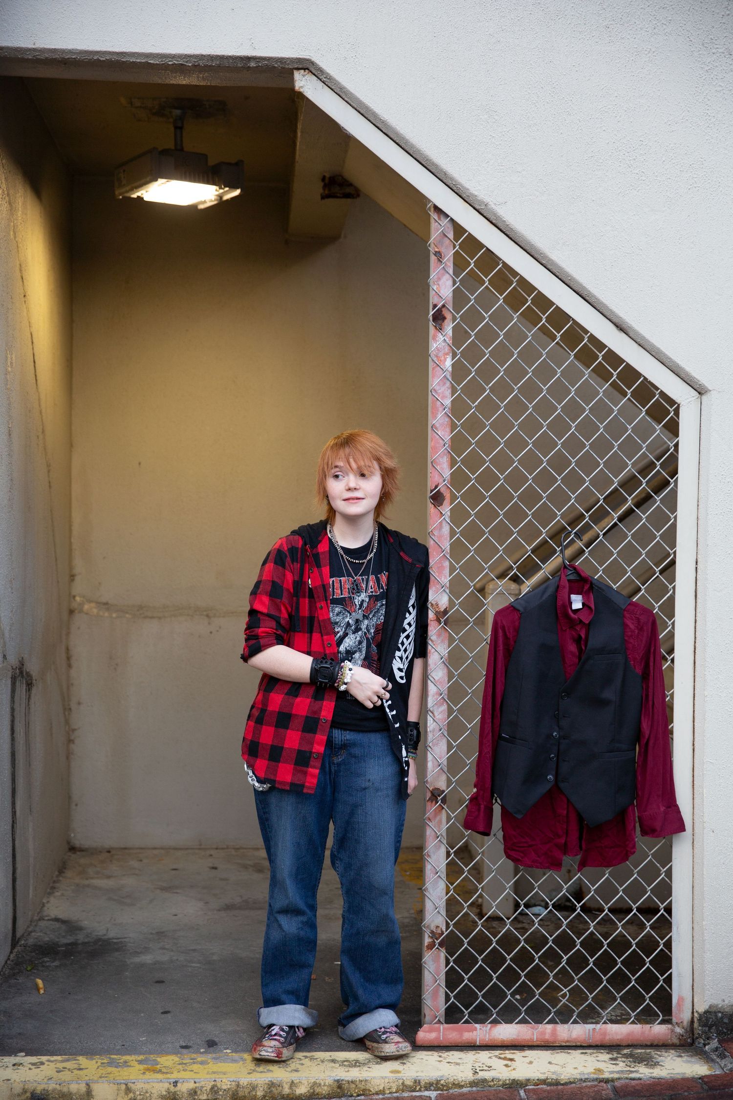 Young person with red hair in plaid shirt stands in doorway with chain-link fence and hanging jacket. Downtown Rome, GA