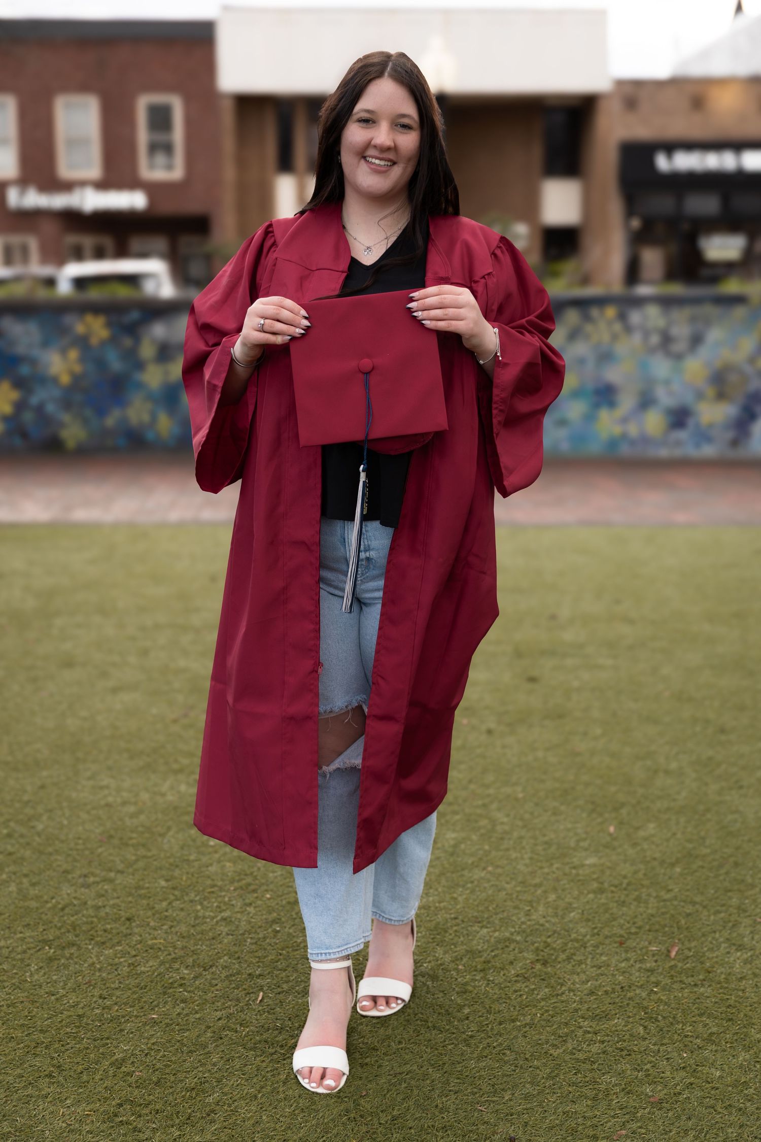 Young woman smiling in red graduation gown and cap on green lawn with shops in background-Northwest Georgia