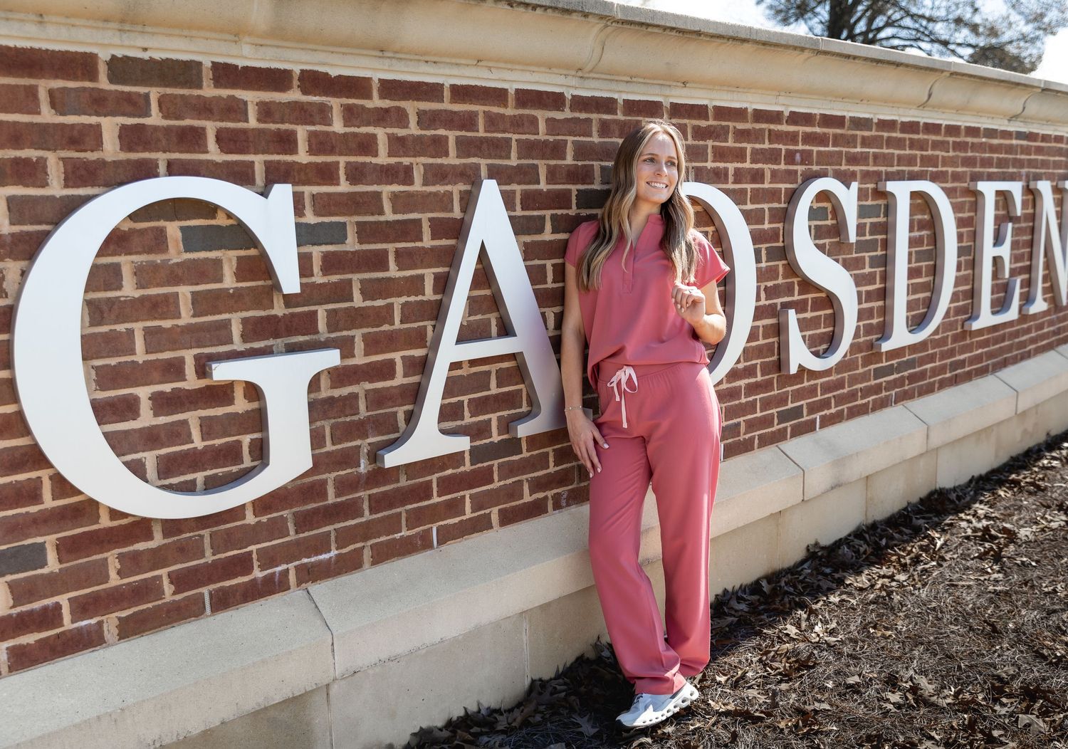 Woman in pink scrubs poses beside a brick Gadsden sign outdoors- Jessica M Cooper Photography LLC