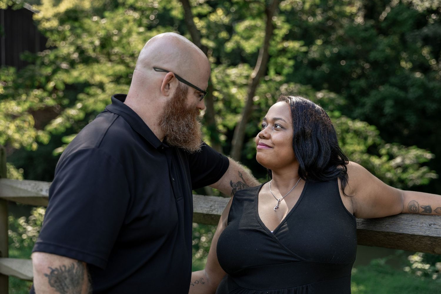 A couple in matching black outfits share a loving gaze while leaning on a wooden fence outdoors. Euharlee Covered Bridge