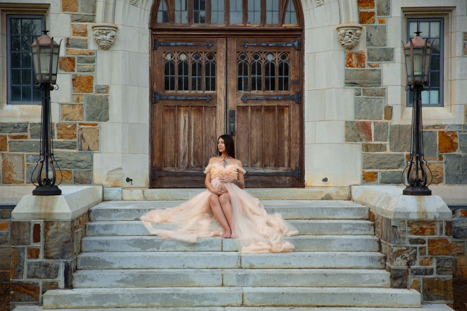 Pregnant woman in elegant blush gown sitting on stone steps before ornate wooden doors of a historic building. Ford Building Berry College