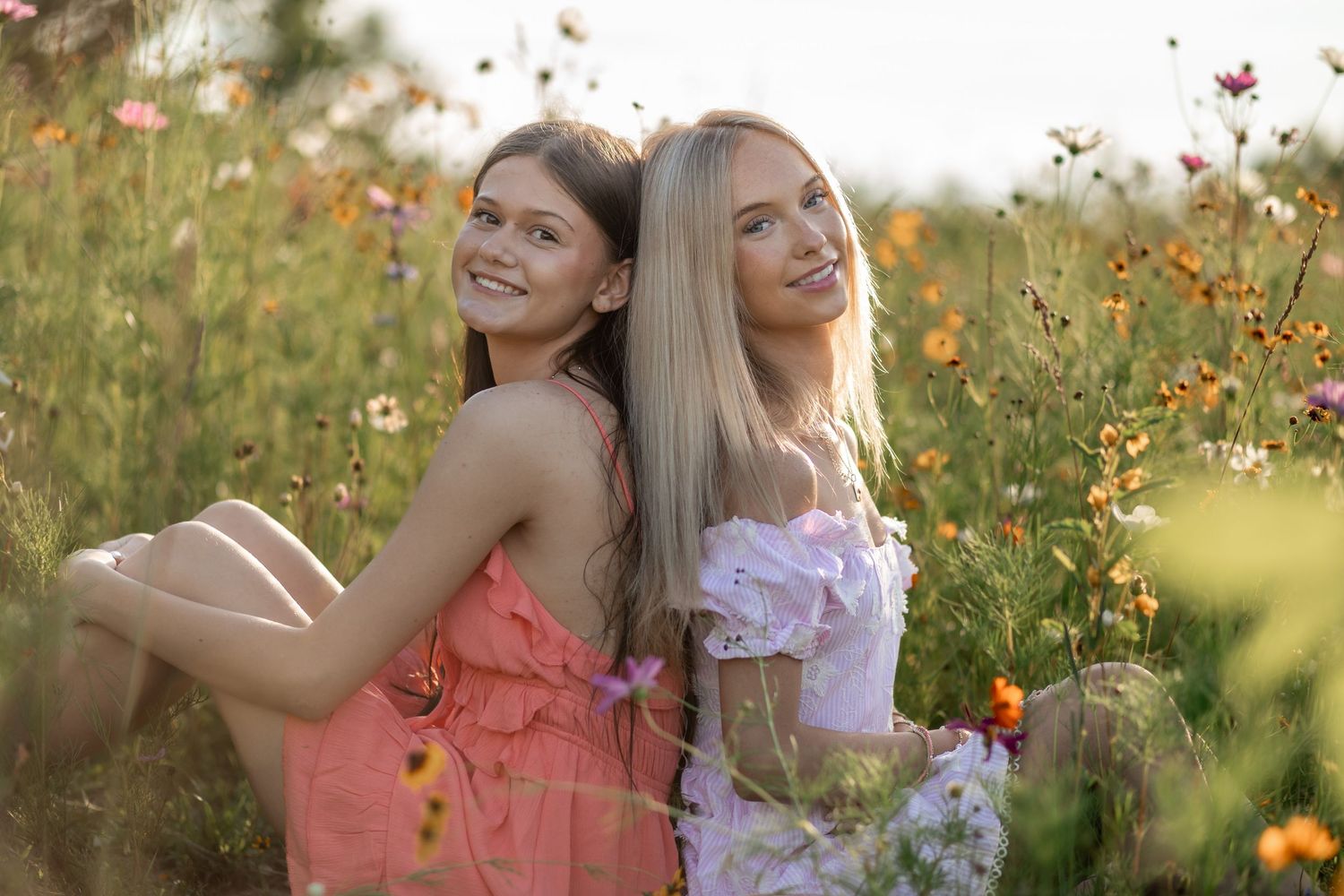Two smiling young women in floral dresses sitting back-to-back in a vibrant wildflower meadow during golden hour.