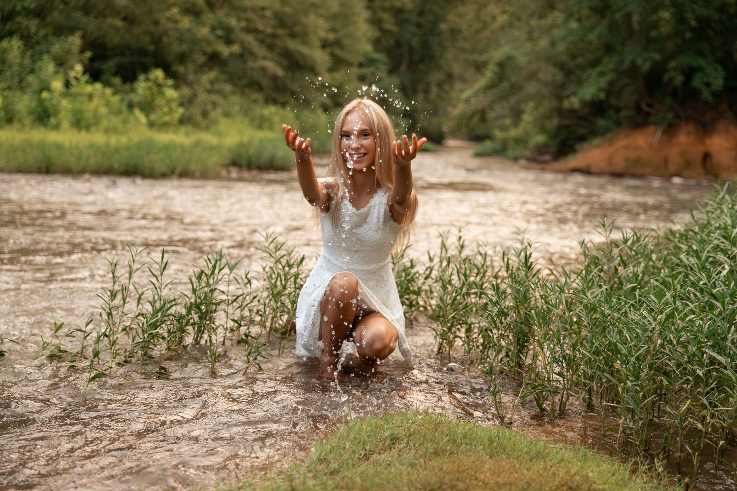 A joyful woman in a white dress splashes water while kneeling in a shallow river surrounded by lush green plants.