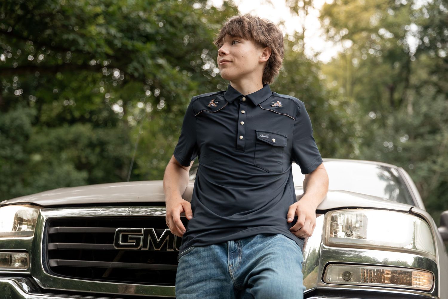 Young man in navy polo leans against a GMC truck outdoors, with lush green trees in the background.