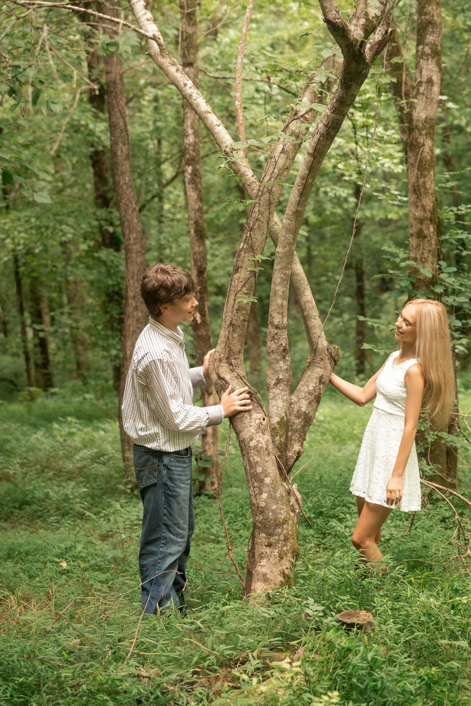 Young couple sharing a romantic moment in a lush green forest, standing near a tree in casual summer attire. Rome, GA