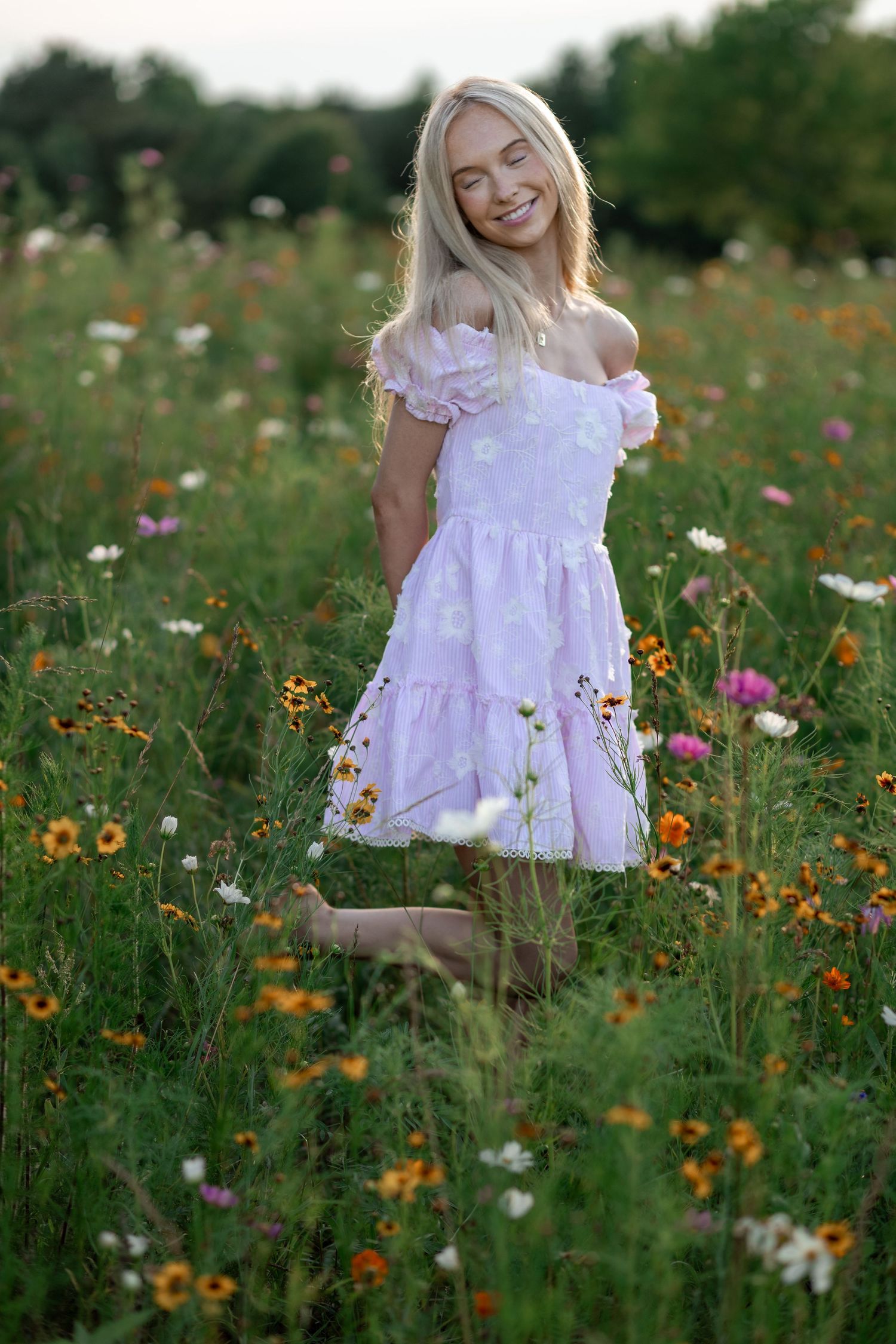 Smiling blonde woman in white off-shoulder dress standing barefoot in a colorful wildflower meadow.