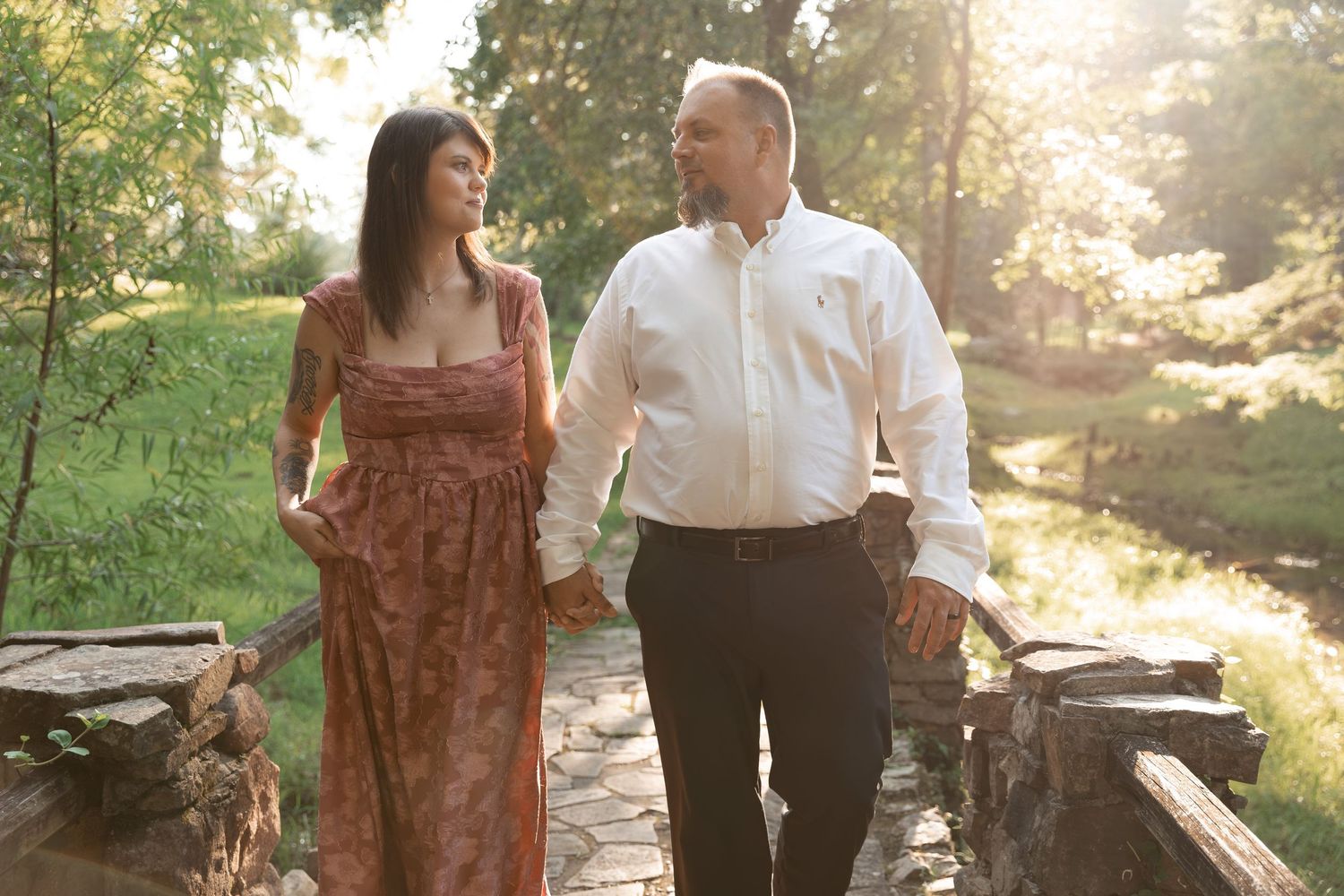 Couple holding hands walking on stone bridge in sunlit park, woman in floral dress, man in white shirt. Berry College, Rome, GA