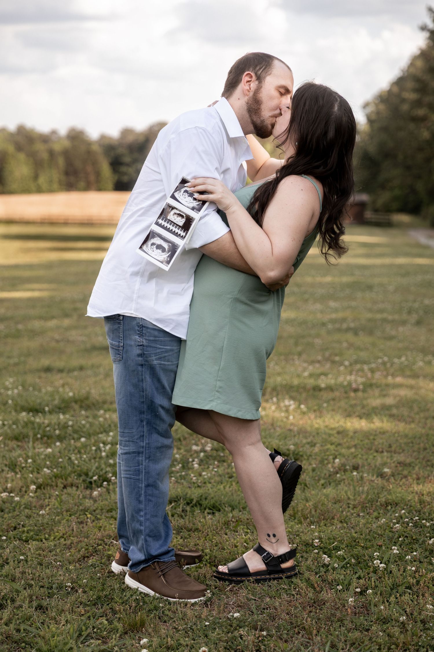 Couple kissing outdoors in a field, holding ultrasound photos, announcing pregnancy in a romantic maternity photo shoot. Berry College