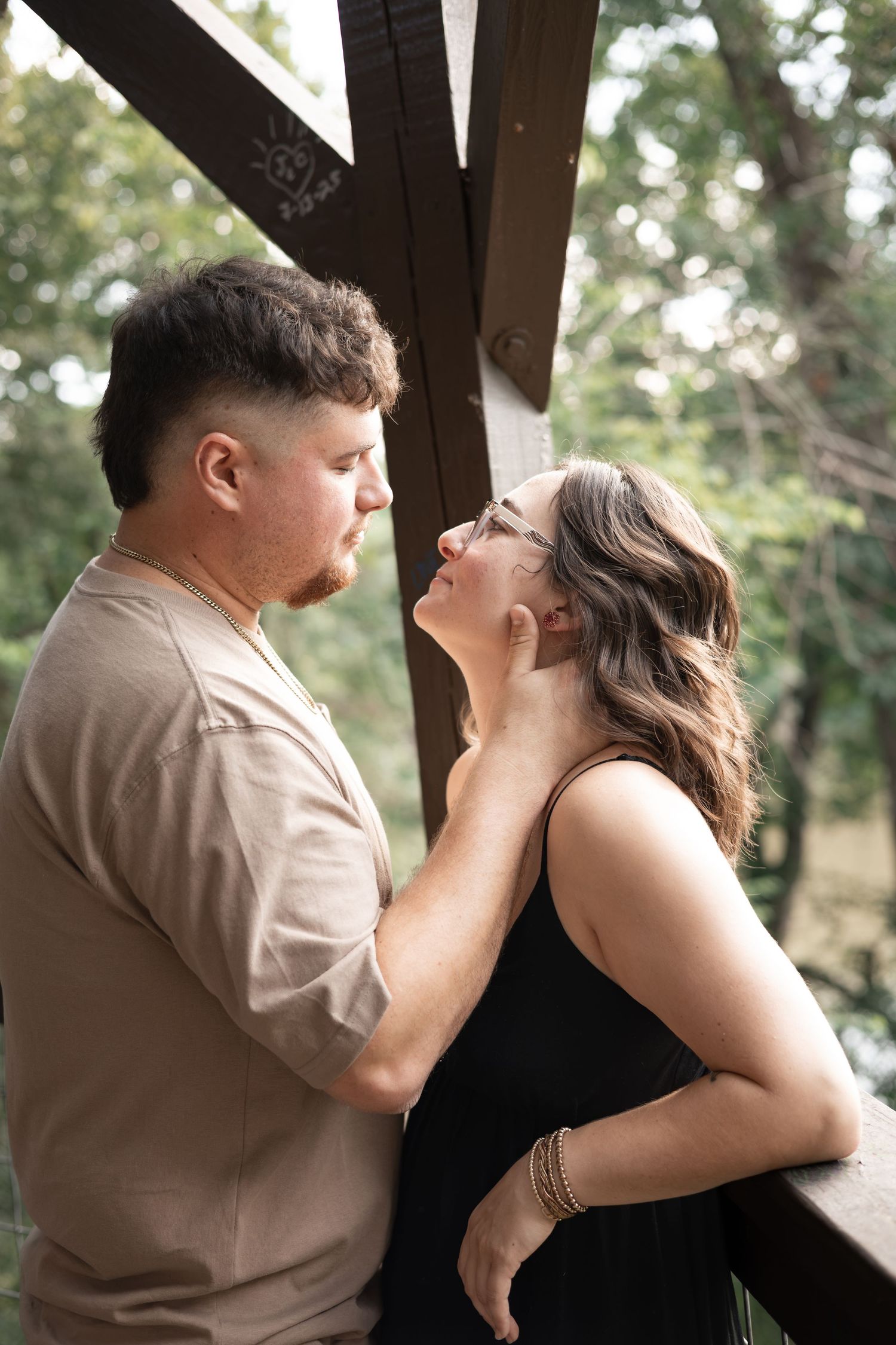 A couple shares a romantic moment on a wooden deck surrounded by lush green trees, man tenderly holding womans face.
