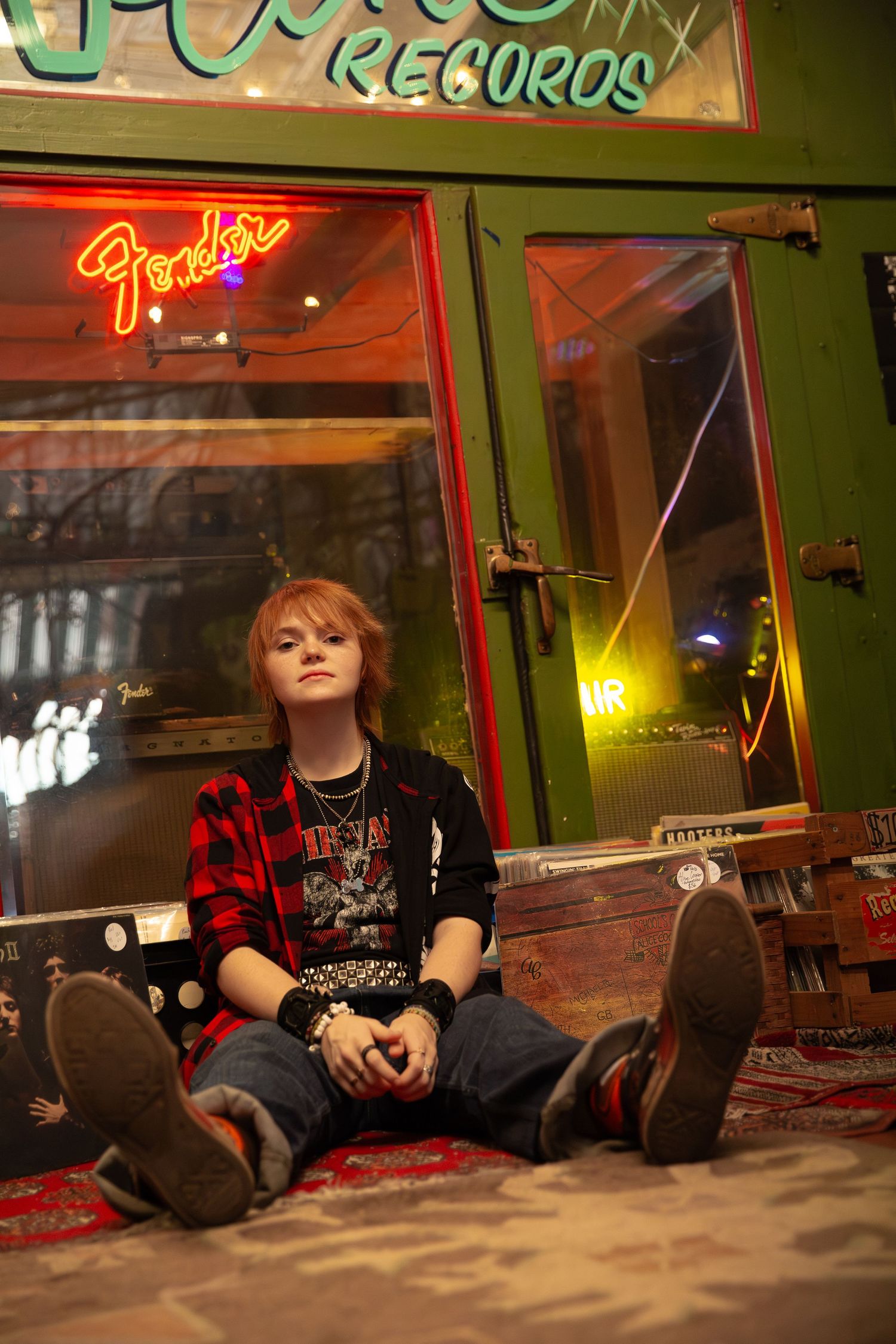 Young person in plaid shirt sitting outside a vintage record store with Fender neon sign- Senior portraits record store downtown Rome, GA