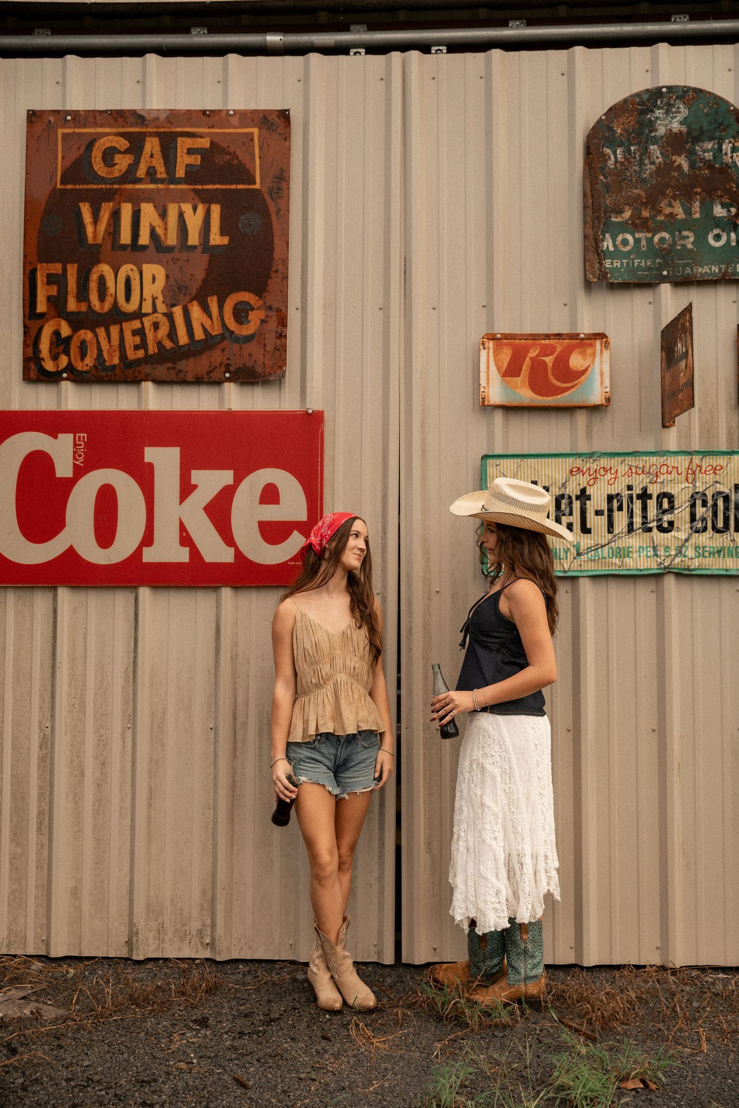 Two women in cowgirl attire chat beside vintage Coke and GAF signs on a rustic corrugated metal wall.