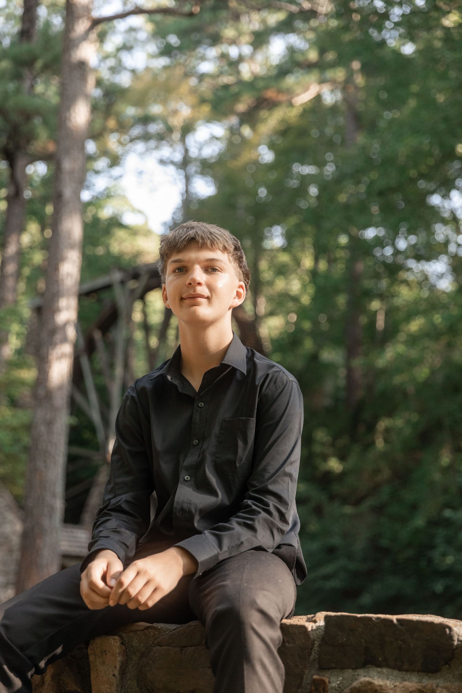 Young man in black shirt sitting on stone wall surrounded by lush green forest trees outdoors.