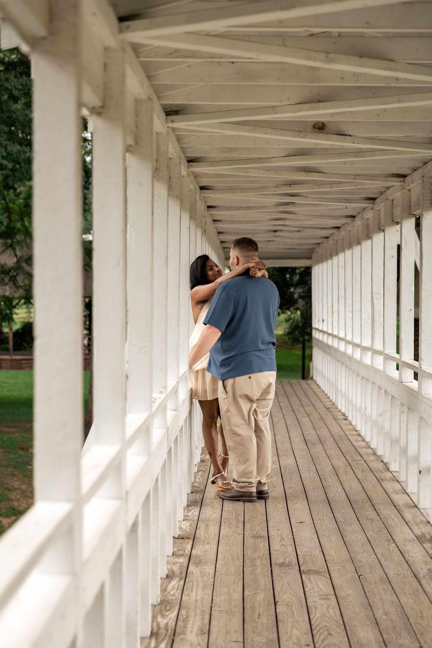 Couple embracing on a white wooden covered bridge walkway surrounded by lush green trees. Rockmart, GA