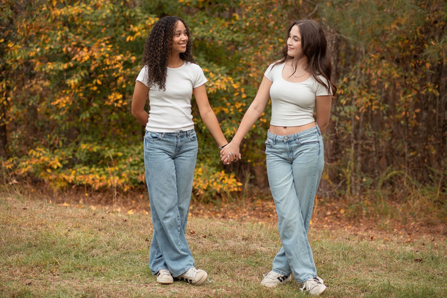 Two friends wearing white tops and jeans hold hands while walking outdoors with autumn foliage in the background- Rome GA