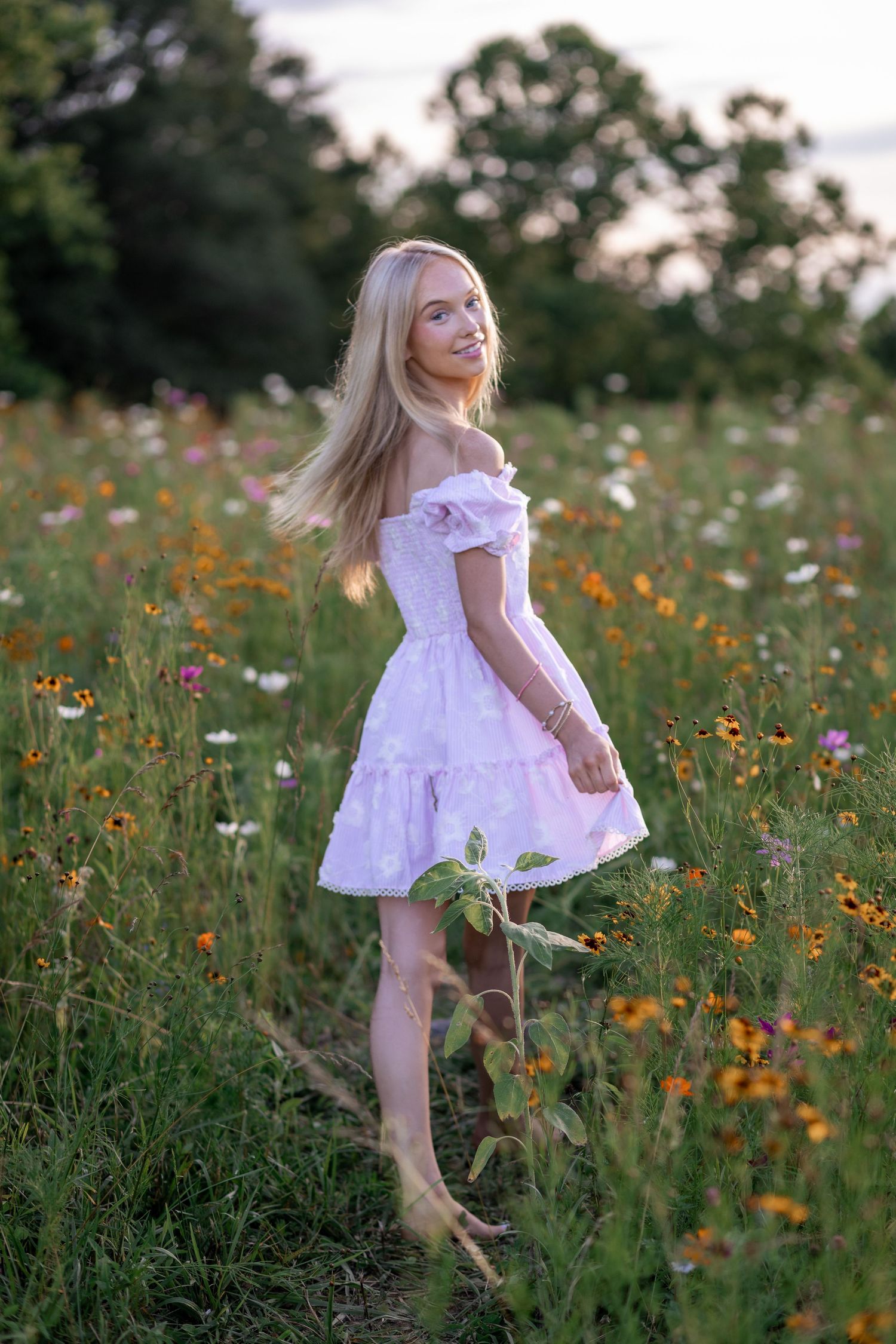 Blonde woman in white off-shoulder dress twirling in a colorful wildflower field at sunset.