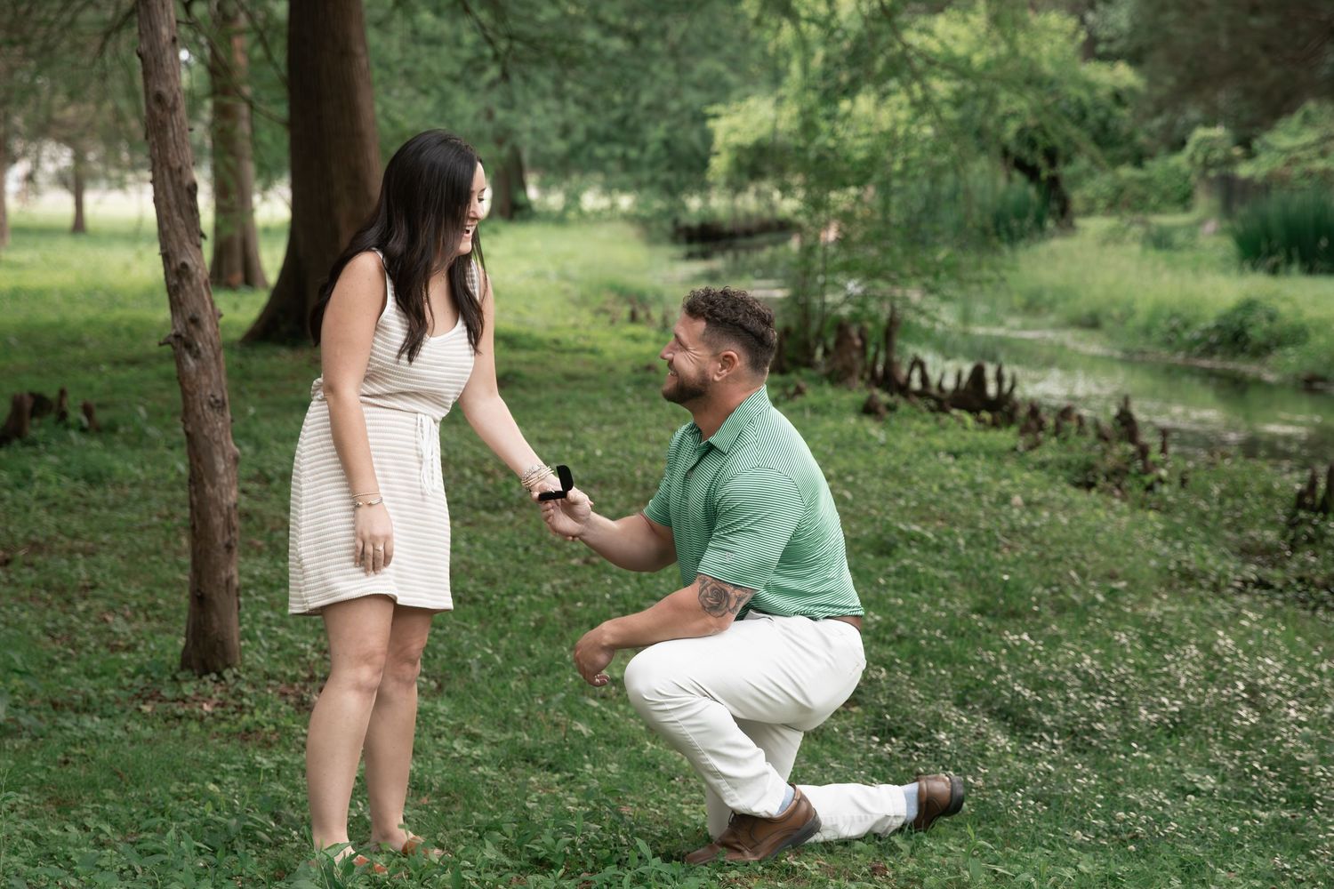 Man kneeling on one knee proposing to woman in white dress outdoors in lush green park setting. Cedartown, GA