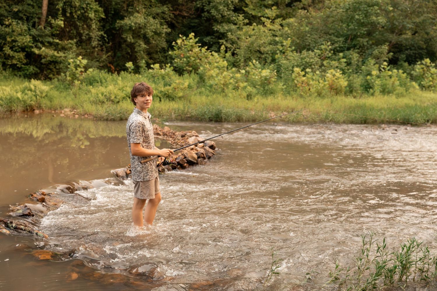Young boy fishing in a shallow rocky river surrounded by lush green trees and grass on a summer day.
