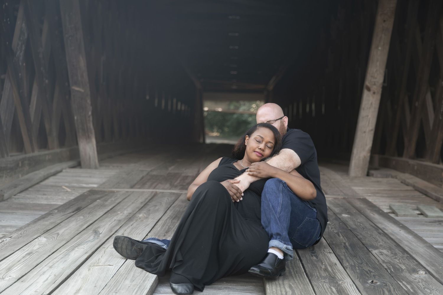 Interracial couple sitting on wooden floor of covered bridge, woman leaning on mans chest in romantic embrace. Euharlee Covered Bridge