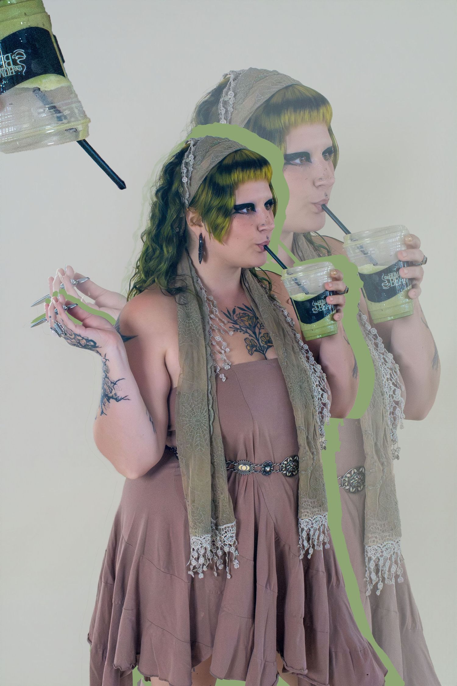 Two women in bohemian mauve dresses with green makeup and scarves drinking iced beverages against white background.