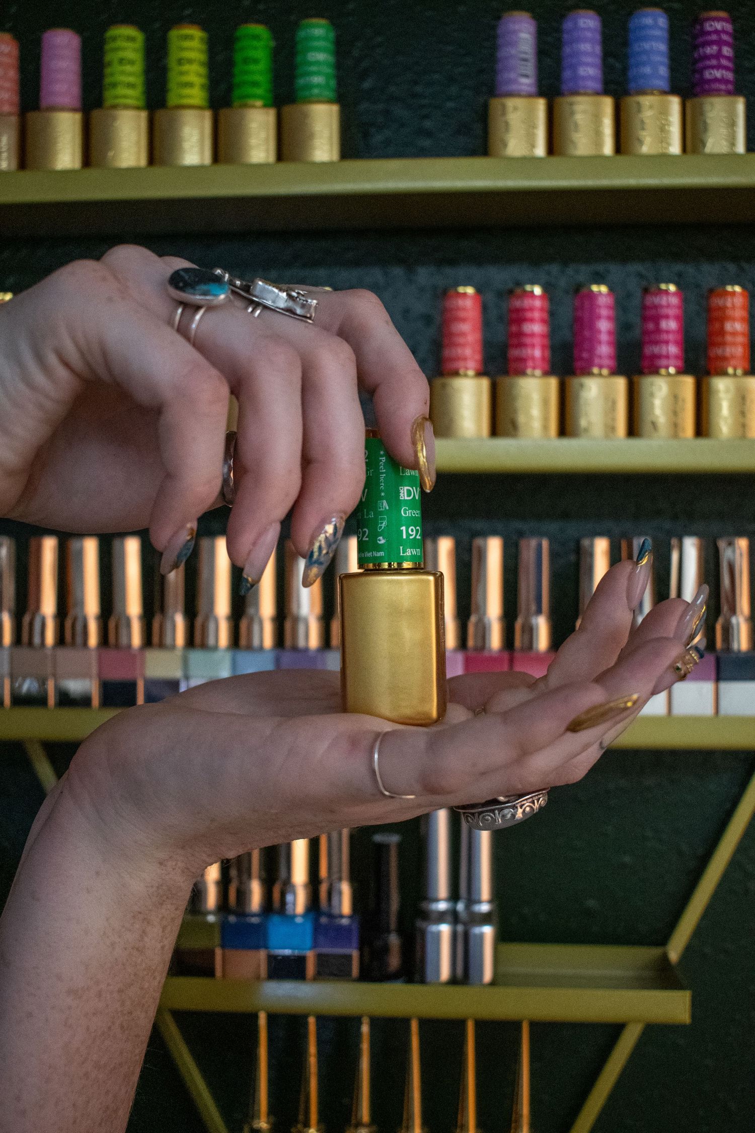 Hands with long nails holding a gold nail polish bottle in front of colorful thread spools on shelves.