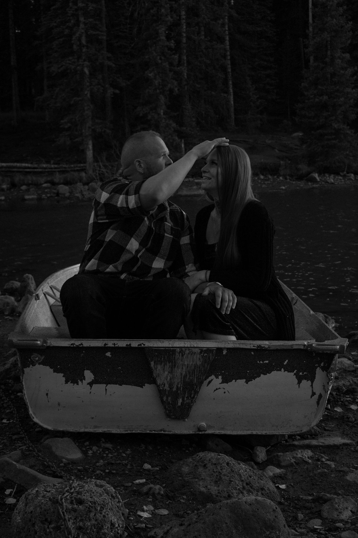Black and white photo of a couple sitting in a rowboat near a forest lake, man gently touching womans hair.