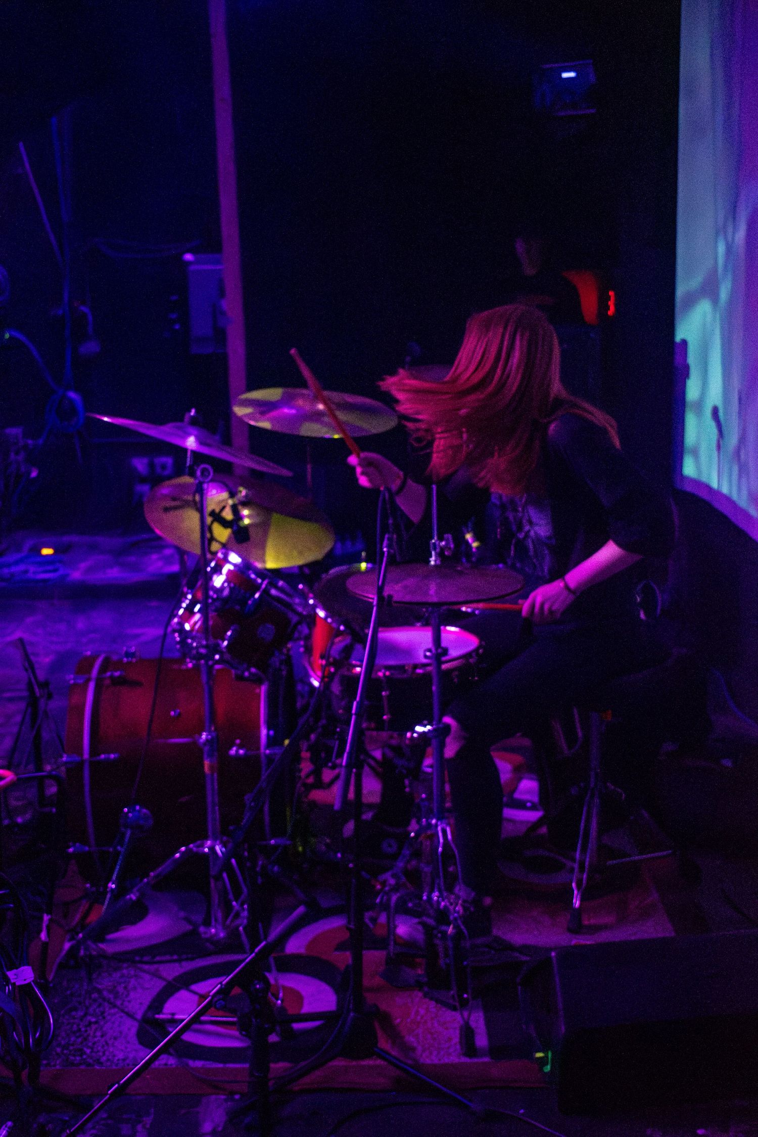 Female drummer with long hair performing on stage under purple lights at a live music concert.