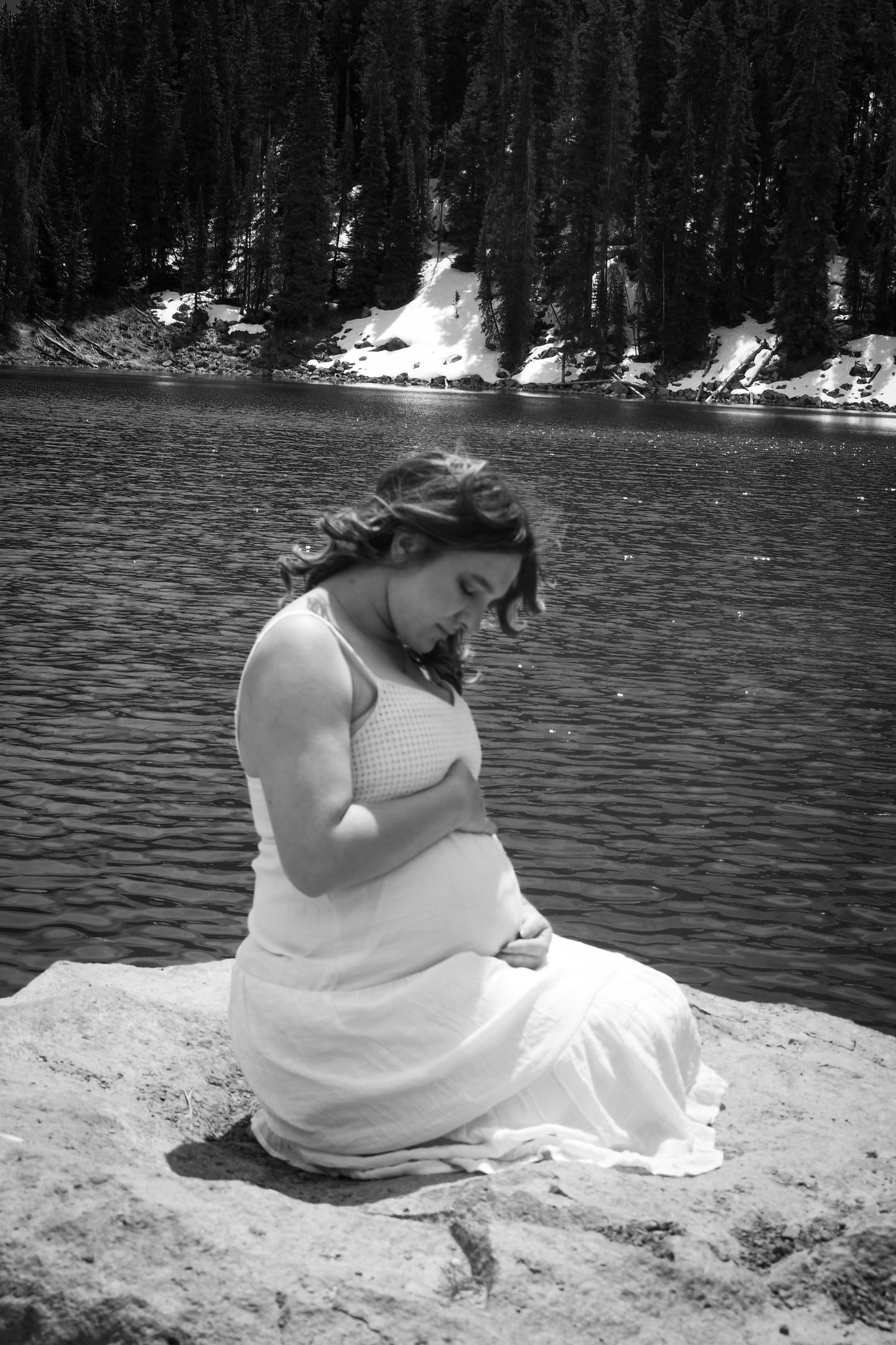 Black and white photo of a pregnant woman sitting on rocks by a mountain lake.