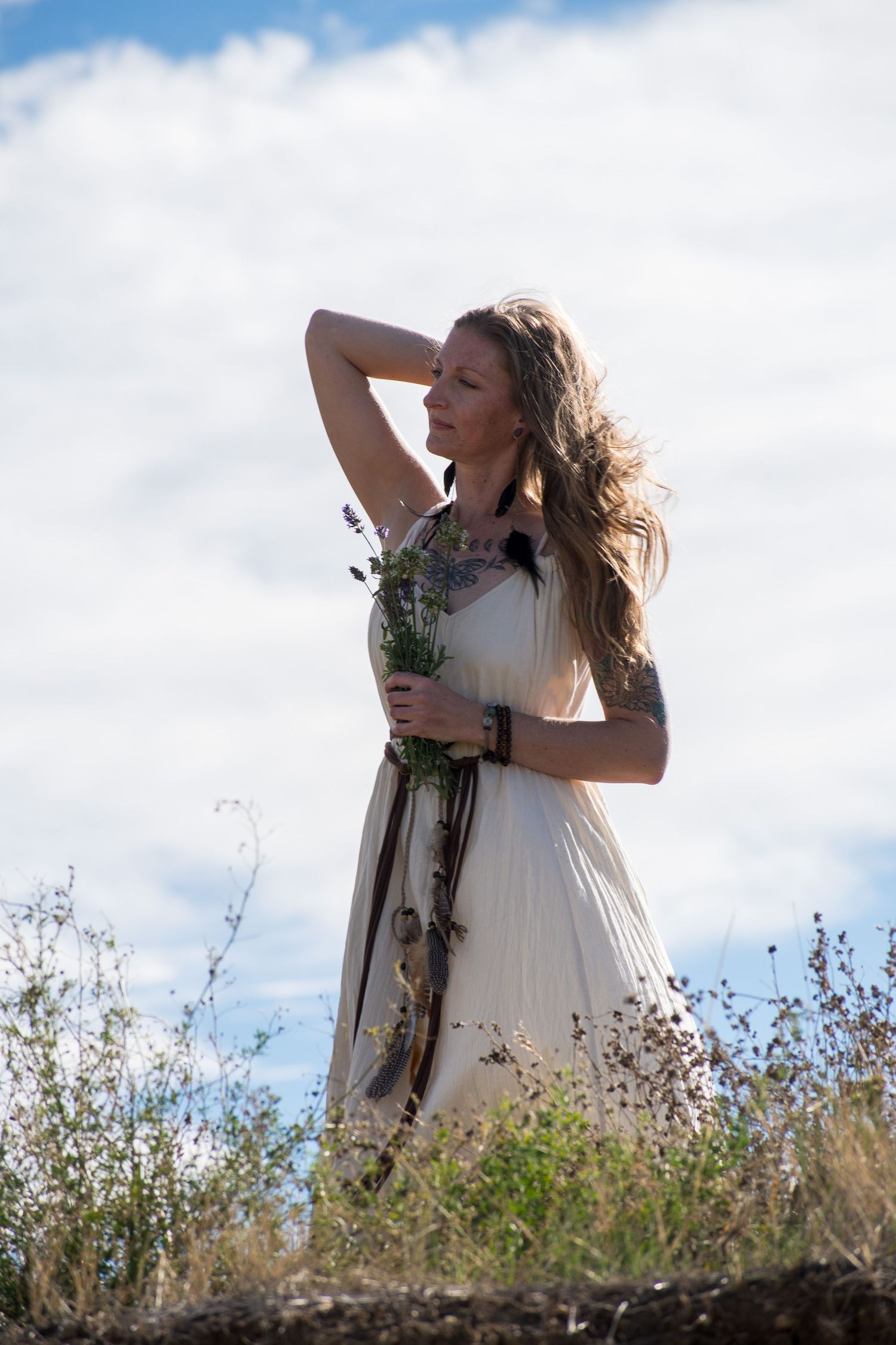 Woman in white dress holding wildflowers in a field, hair blowing in the wind against a cloudy blue sky.