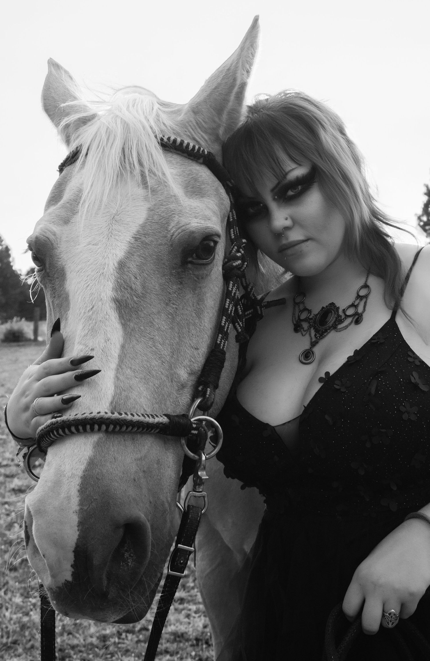 Black and white photo of a gothic woman in black lace dress posing with a white horse outdoors.