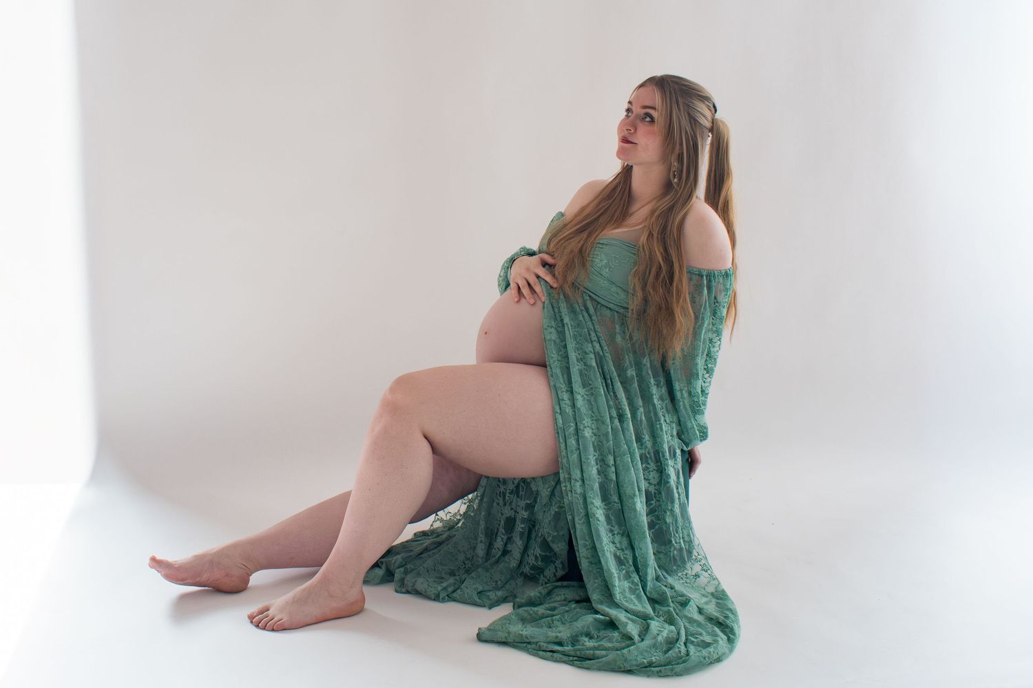 Pregnant woman in flowing teal maternity gown sitting gracefully on white studio floor during maternity photoshoot.