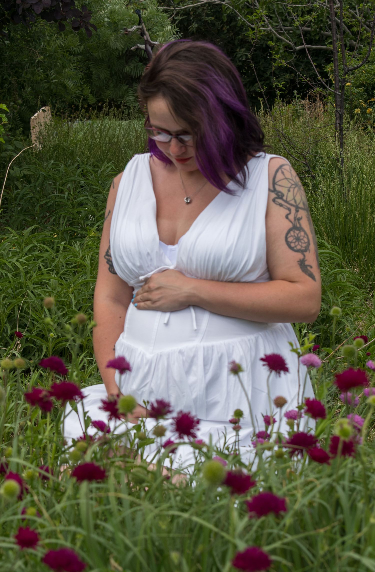 Woman with purple hair and tattoo in white dress among red flowers in a lush green garden.