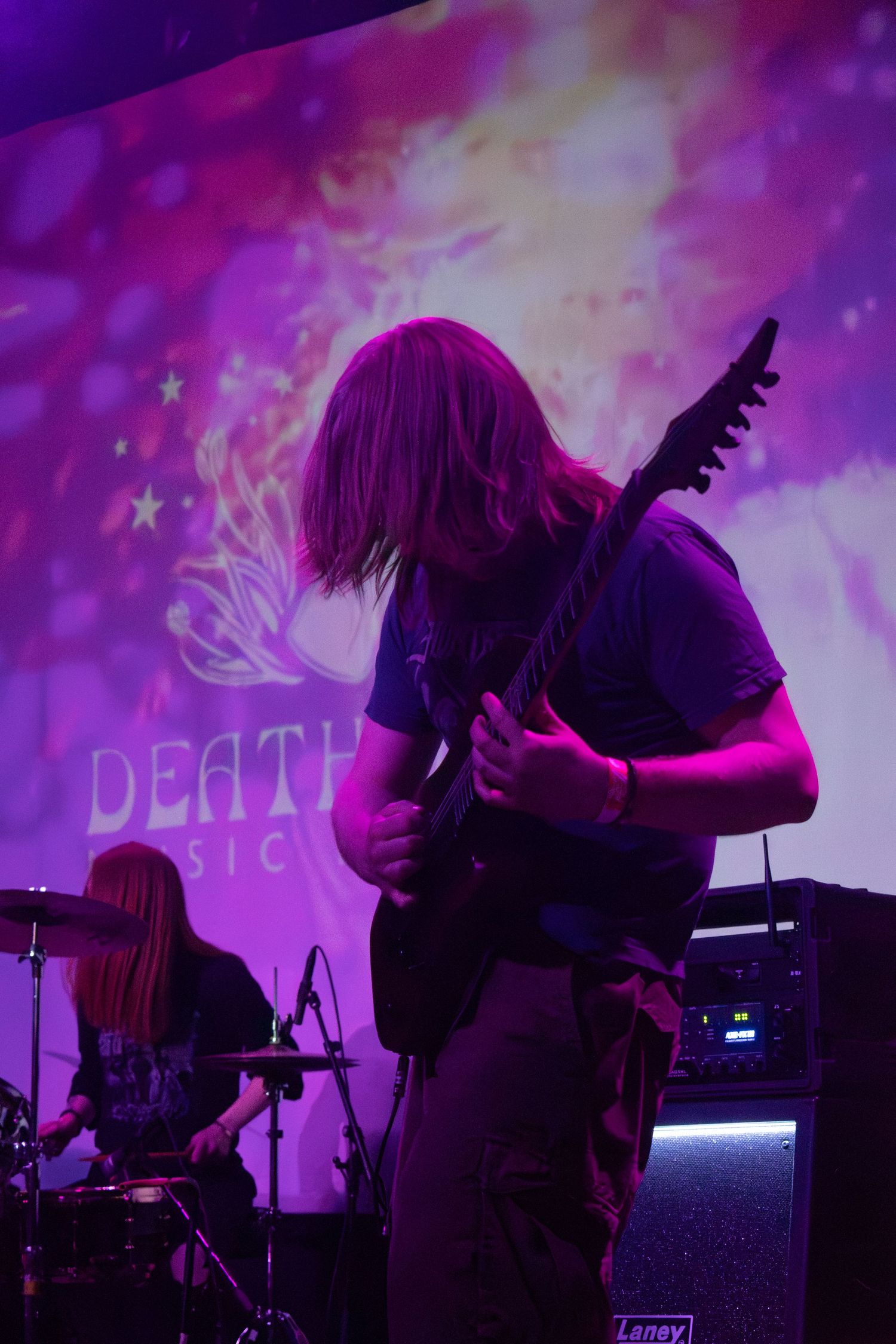 Guitarist performing on stage under purple lights with a drummer and projected backdrop at a Death Music event.