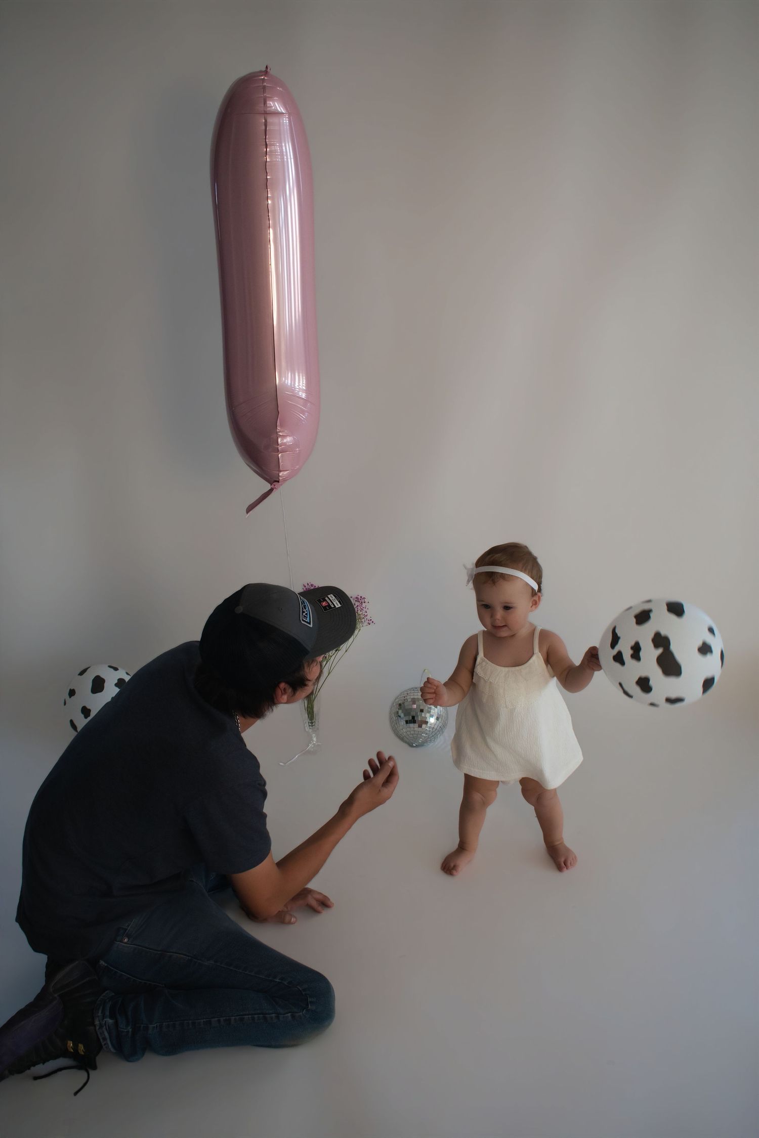 Father kneeling and playing with baby daughter holding balloons during first birthday photo session.