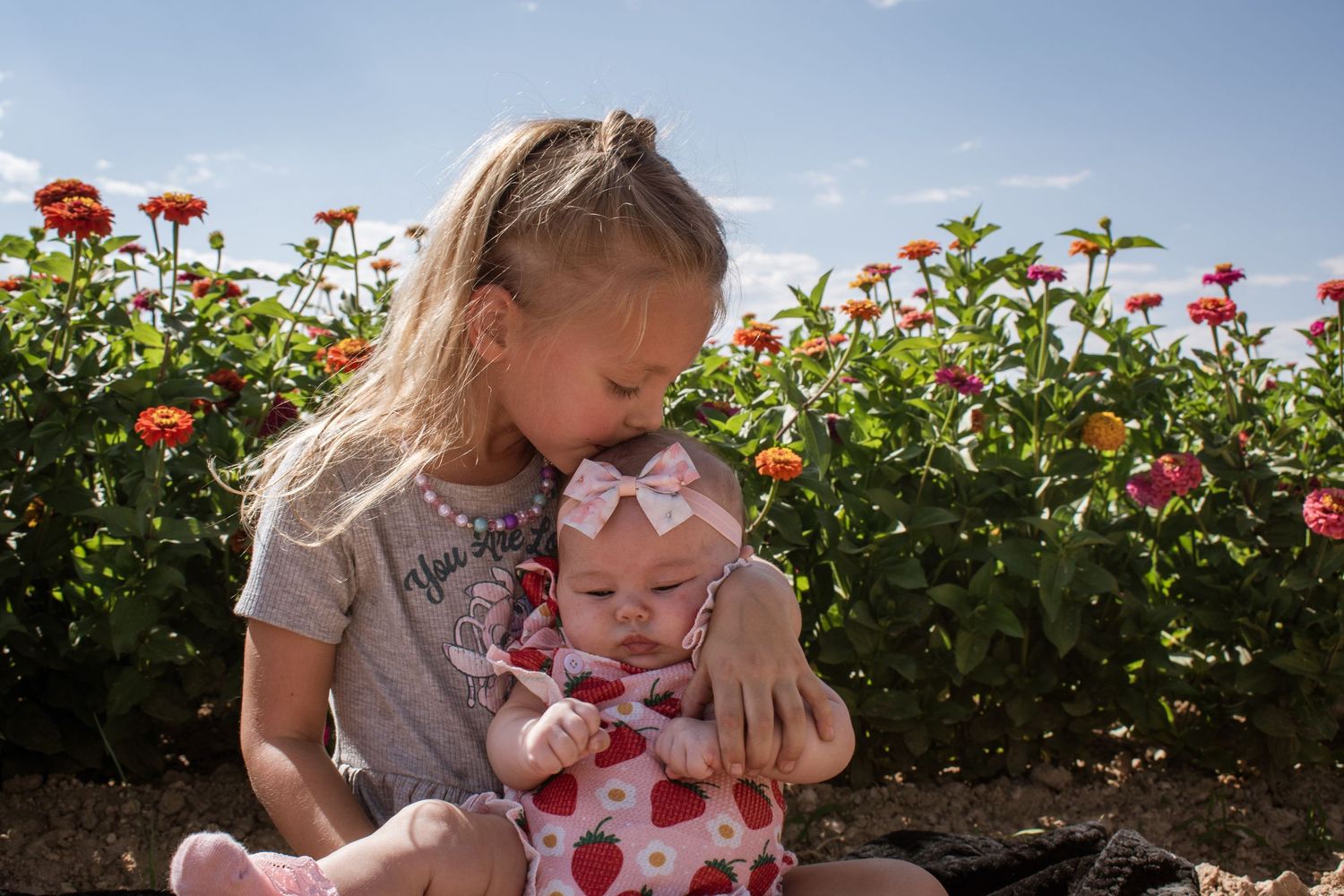 Older sister lovingly kissing baby sibling while sitting in front of colorful flower garden.