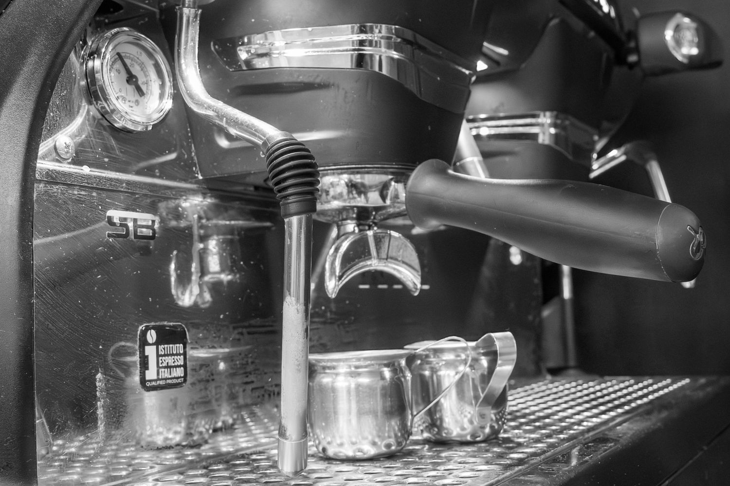 Black and white close-up of a professional espresso machine with steam wand and metal milk pitcher.