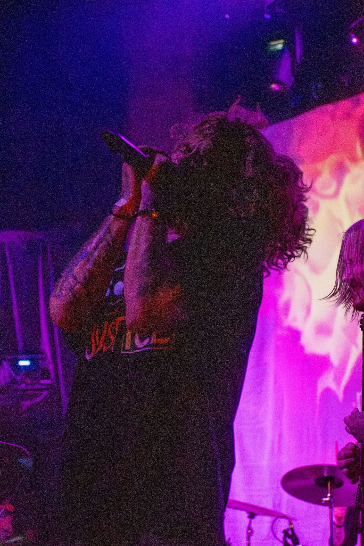 Tattooed rock singer performing on stage with microphone, purple lighting and flame backdrop at live concert.