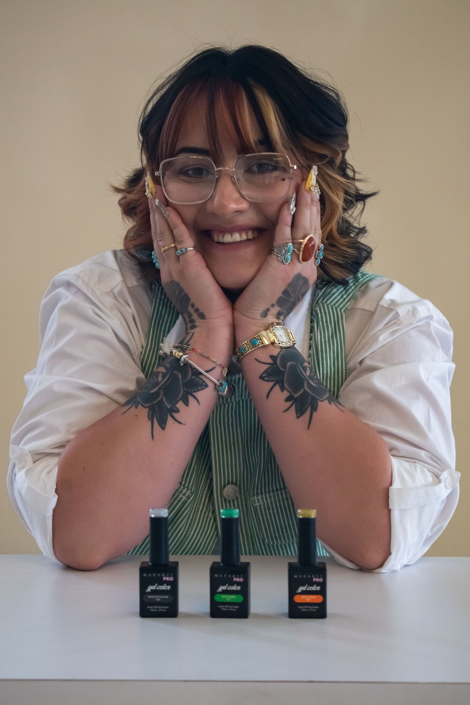 Smiling woman with tattoos and glasses poses with nail polish bottles on white table.
