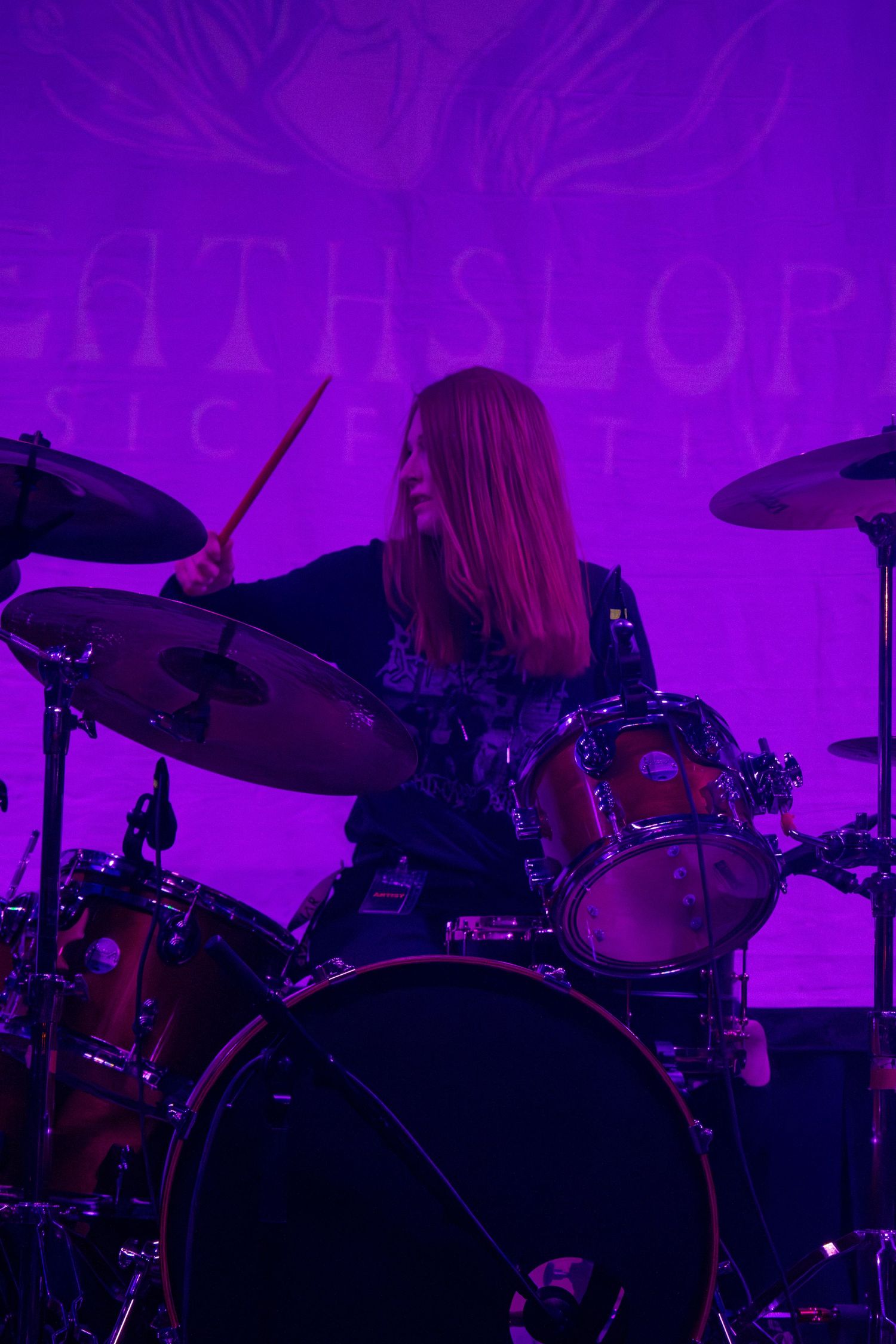 Female drummer performing on stage under purple lights at a music festival, playing drums with sticks.