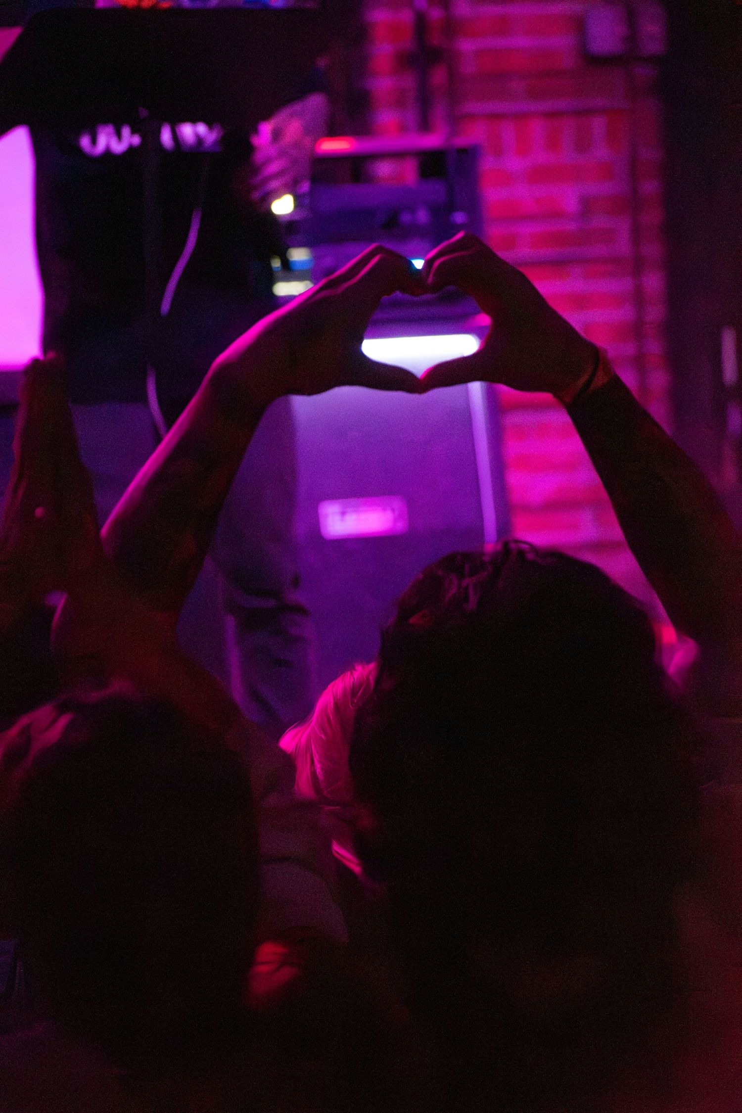 Person making a heart shape with hands at a concert with pink and purple neon lights in the background.