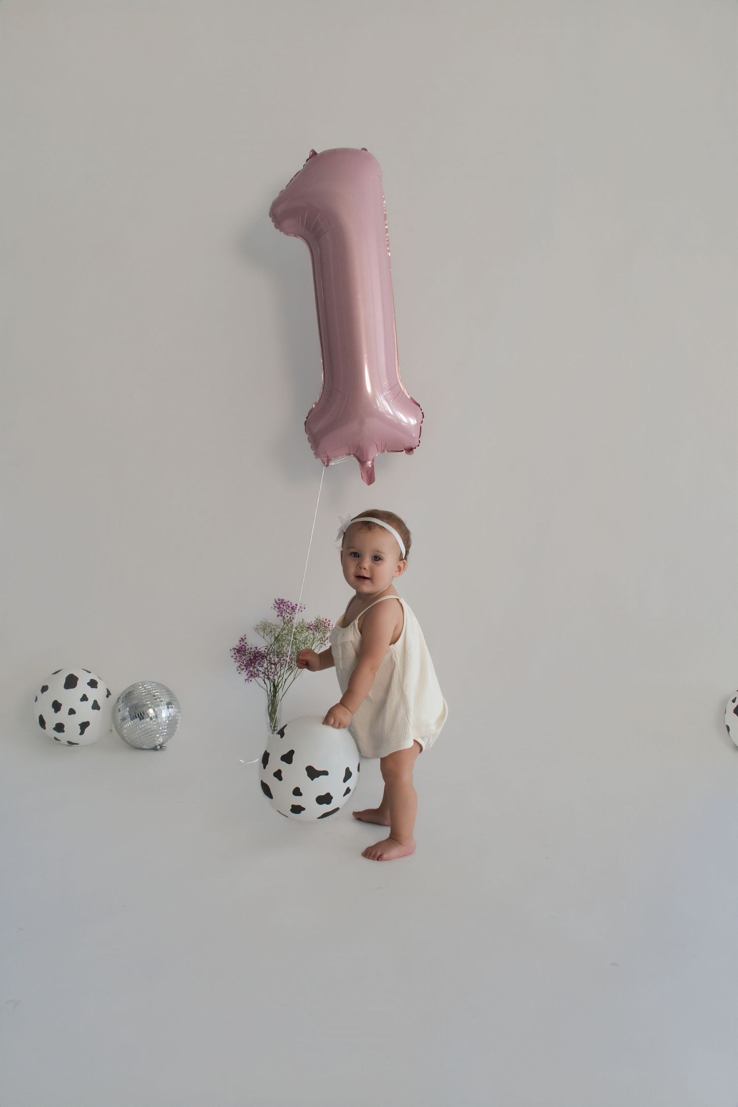 Happy baby girl standing next to a pink number one balloon celebrating her first birthday.