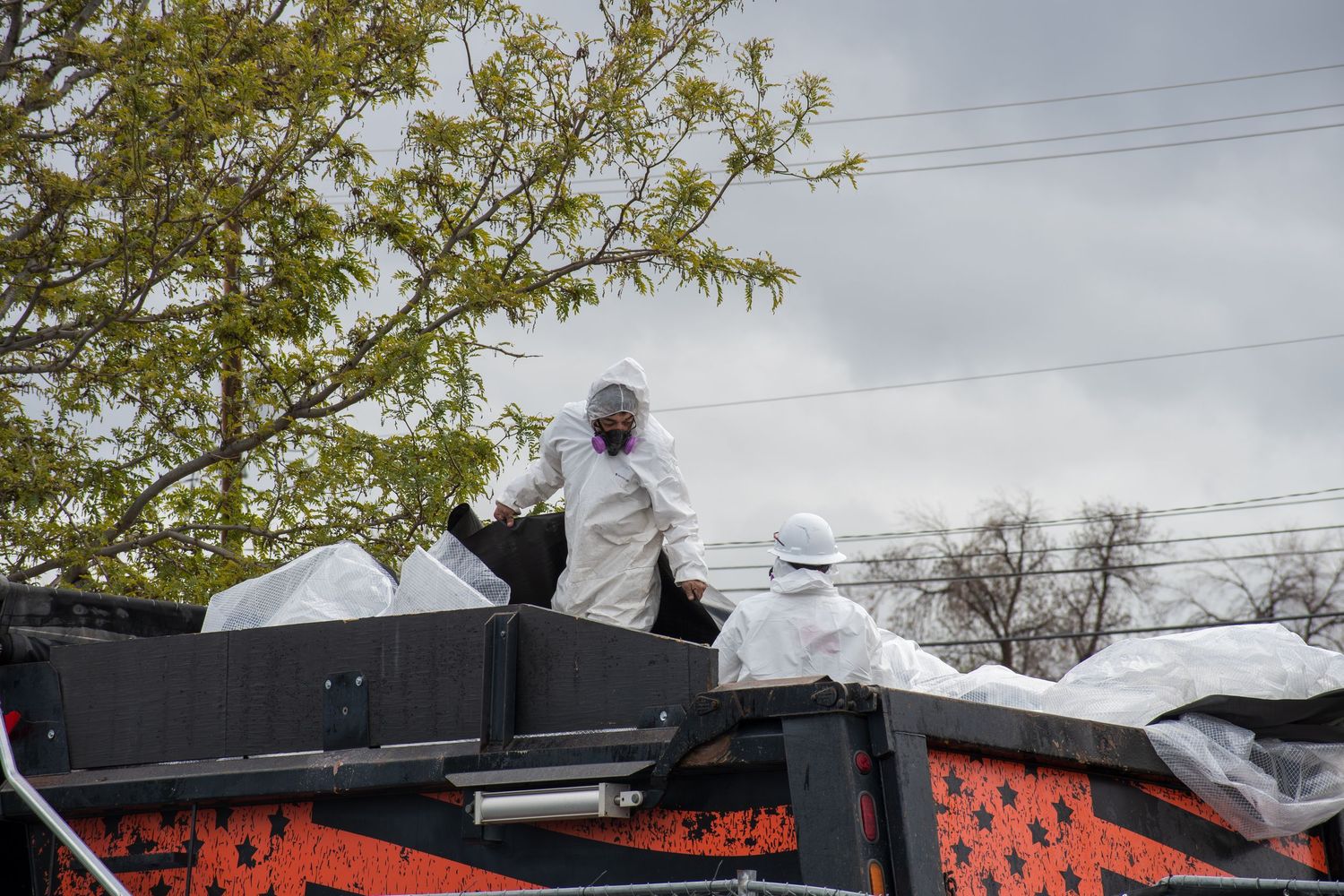 Workers in white hazmat suits handle white body bags on a truck bed outdoors.