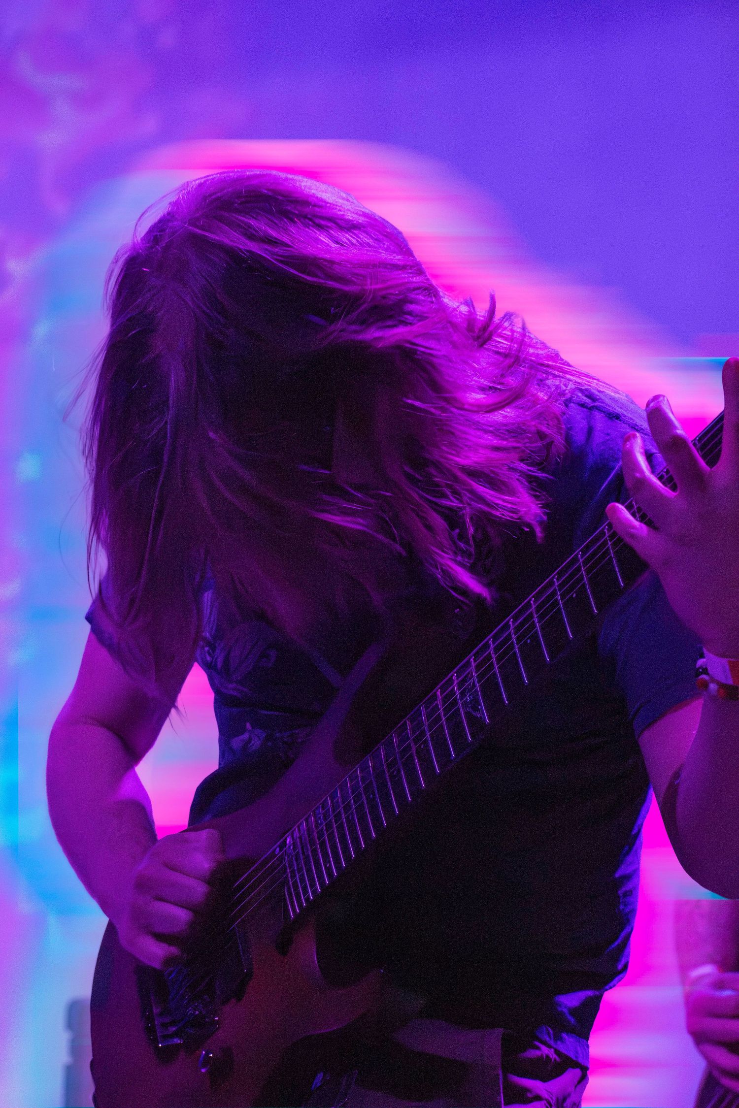 Guitarist headbanging on stage under vibrant purple and pink lights during a live rock concert performance.