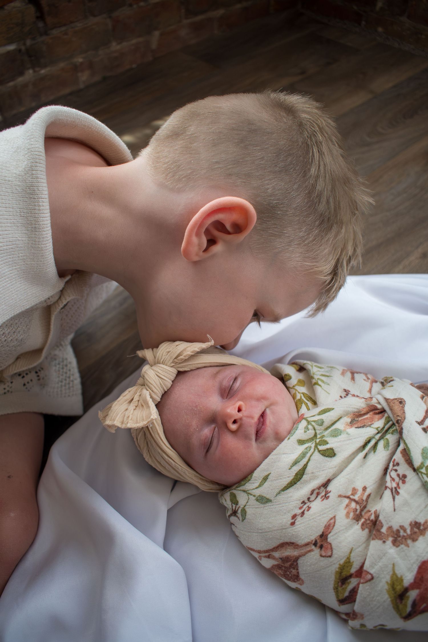Older boy gently kissing forehead of sleeping newborn baby girl swaddled in floral blanket with bow headband.