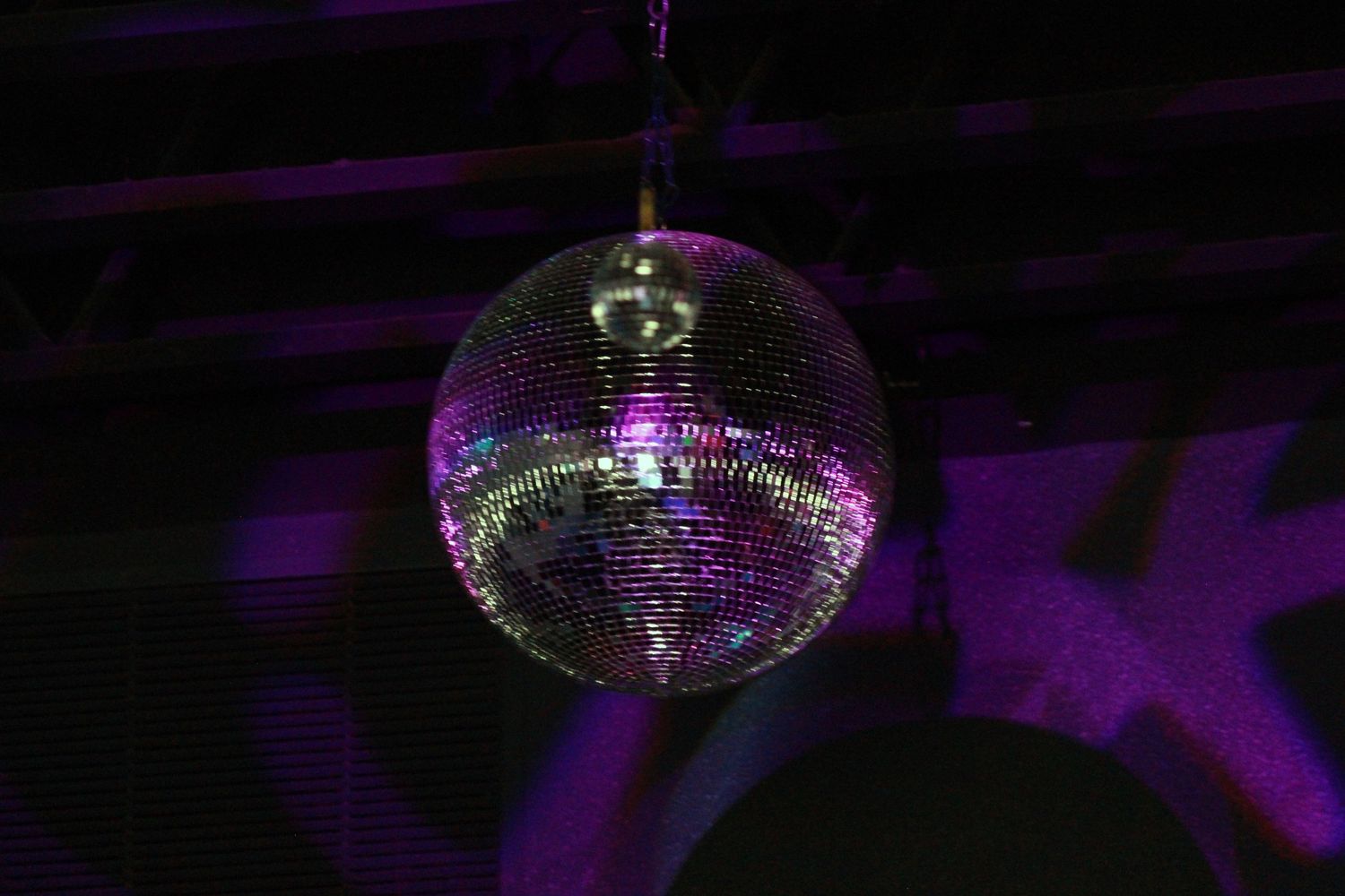 A shimmering disco ball hanging from a ceiling, reflecting purple and green lights in a dark nightclub.