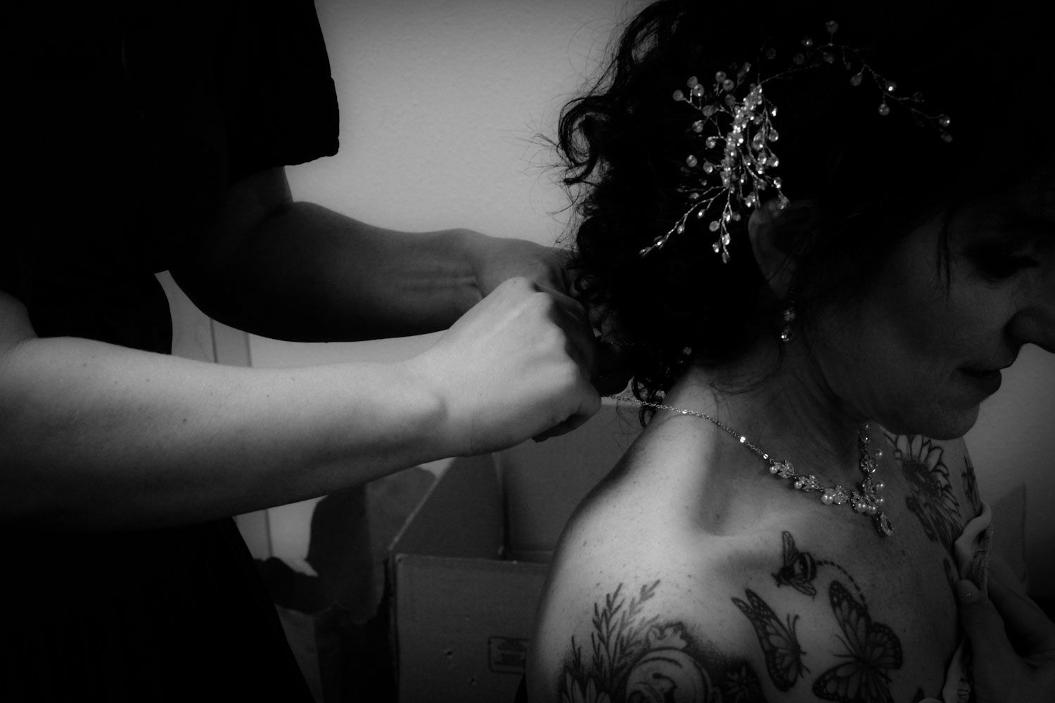 Black and white close-up of a tattooed bride having her necklace fastened before her wedding ceremony.