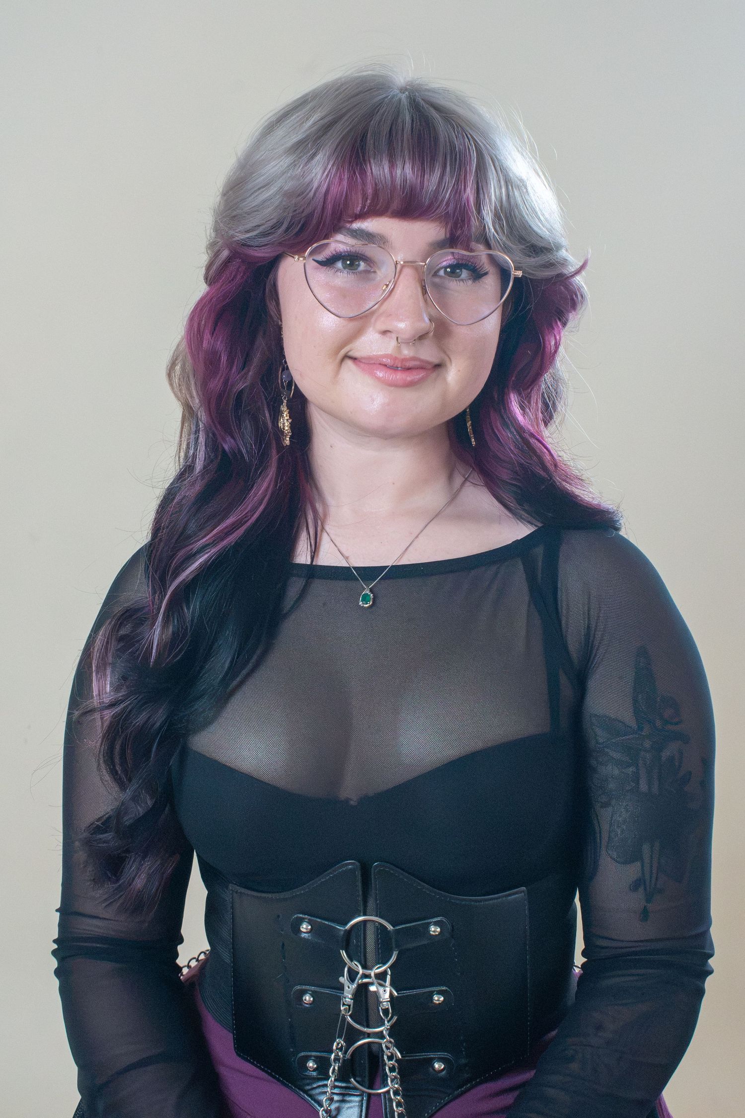Young woman with purple and silver hair, glasses, black mesh top and corset, smiling at camera.