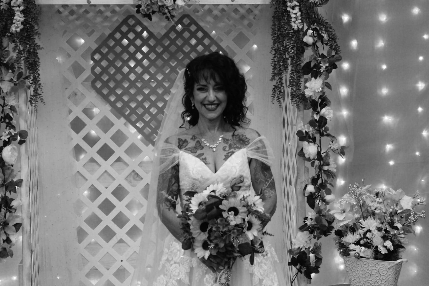 Smiling bride in white gown holds bouquet standing before floral lattice backdrop with lights.